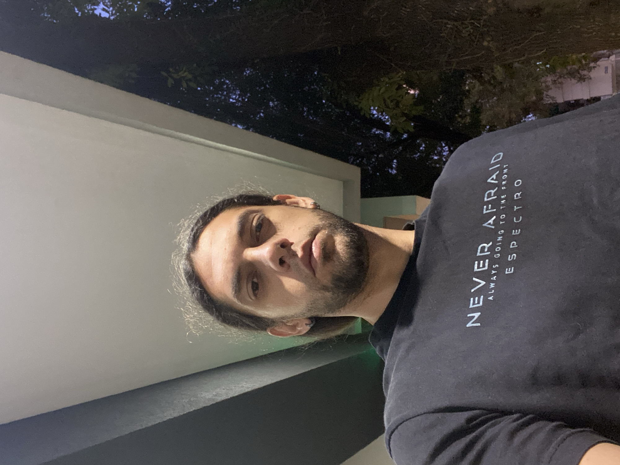 A low-angle selfie captures a male individual at night. The man, appearing to be in his late 20s to early 30s, has dark, long, wavy hair pulled back, a short beard, and a neutral expression. He wears a dark navy blue or black short-sleeved t-shirt with the light-colored text "NEVER AFRAID" in large capital letters, "ALWAYS GOING TO THE FRONT" in smaller capital letters below, and "ESPECTRO" in small capital letters at the bottom. A single silver-colored hoop earring is visible in his left ear. Harsh, direct artificial lighting from above illuminates the subject's face and the immediate background. Overhead, an architectural element consists of a light grey or white angled canopy or overhang, flanked on the far left by a darker grey or black vertical panel. A faint green light source is visible behind the subject's head on the left. Beyond the canopy, on the right side of the frame, dark tree canopies are silhouetted against a deep indigo to purplish-blue night sky, indicating an outdoor or semi-outdoor setting. The specific location within Buenos Aires, Argentina, cannot be determined from the visual details present in the image.