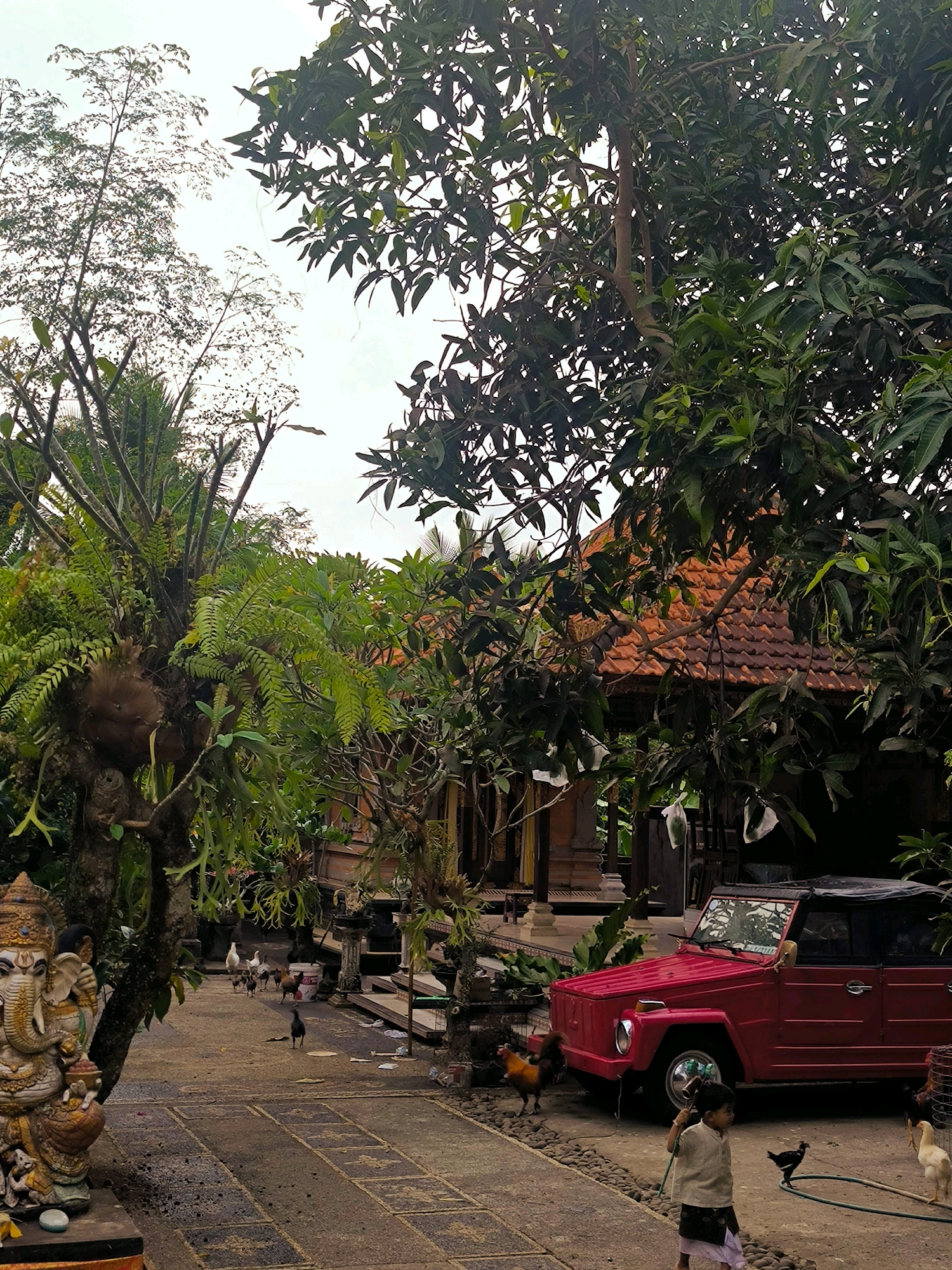 In Selemadeg, Indonesia, a bright red vintage Volkswagen Type 181, also known as a "Thing," is parked on a paved outdoor area. A young child, wearing a light-colored shirt and a dark patterned sarong, walks towards the vehicle, holding a green hose. Several chickens are scattered around the scene, including one near the child and another to the right of the car. In the background, traditional Balinese architecture with red tile roofs is visible, surrounded by lush green trees and ferns. To the left, a large, ornate statue of the Hindu deity Ganesha stands prominently. The ground is a mix of paved sections and small stones. The sky is overcast.