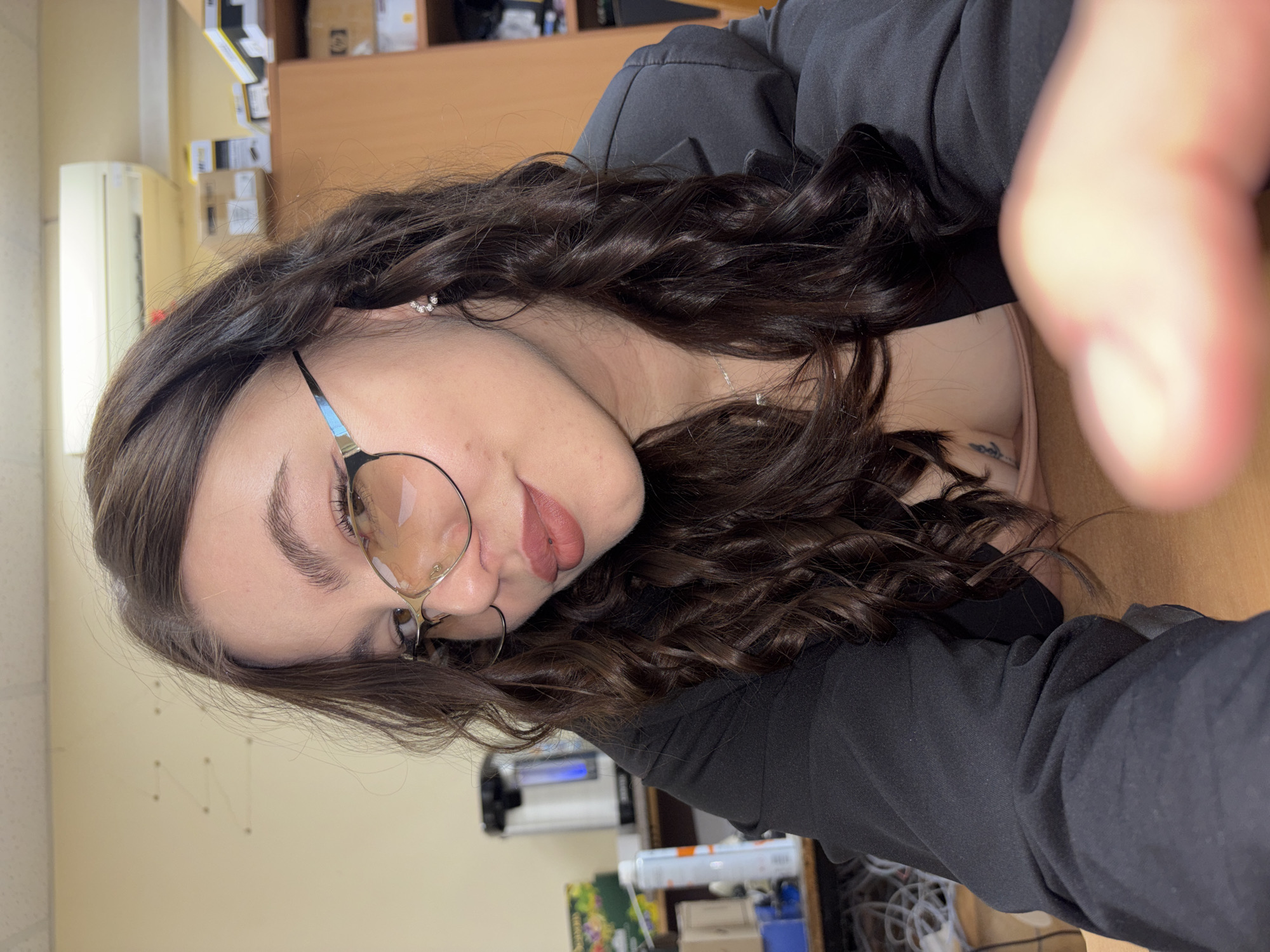 A woman is taking a selfie indoors. She has dark brown curly hair, fair skin, and is wearing round glasses with metal frames. She is wearing a black jacket. A partial view of her collarbone reveals a necklace and a small tattoo.
The background appears to be an office or workspace. There is a beige wall with what appears to be a grid of wires or tacks. A desk and various office supplies are visible.
Based on the tags, the location is in Perm, Russia.