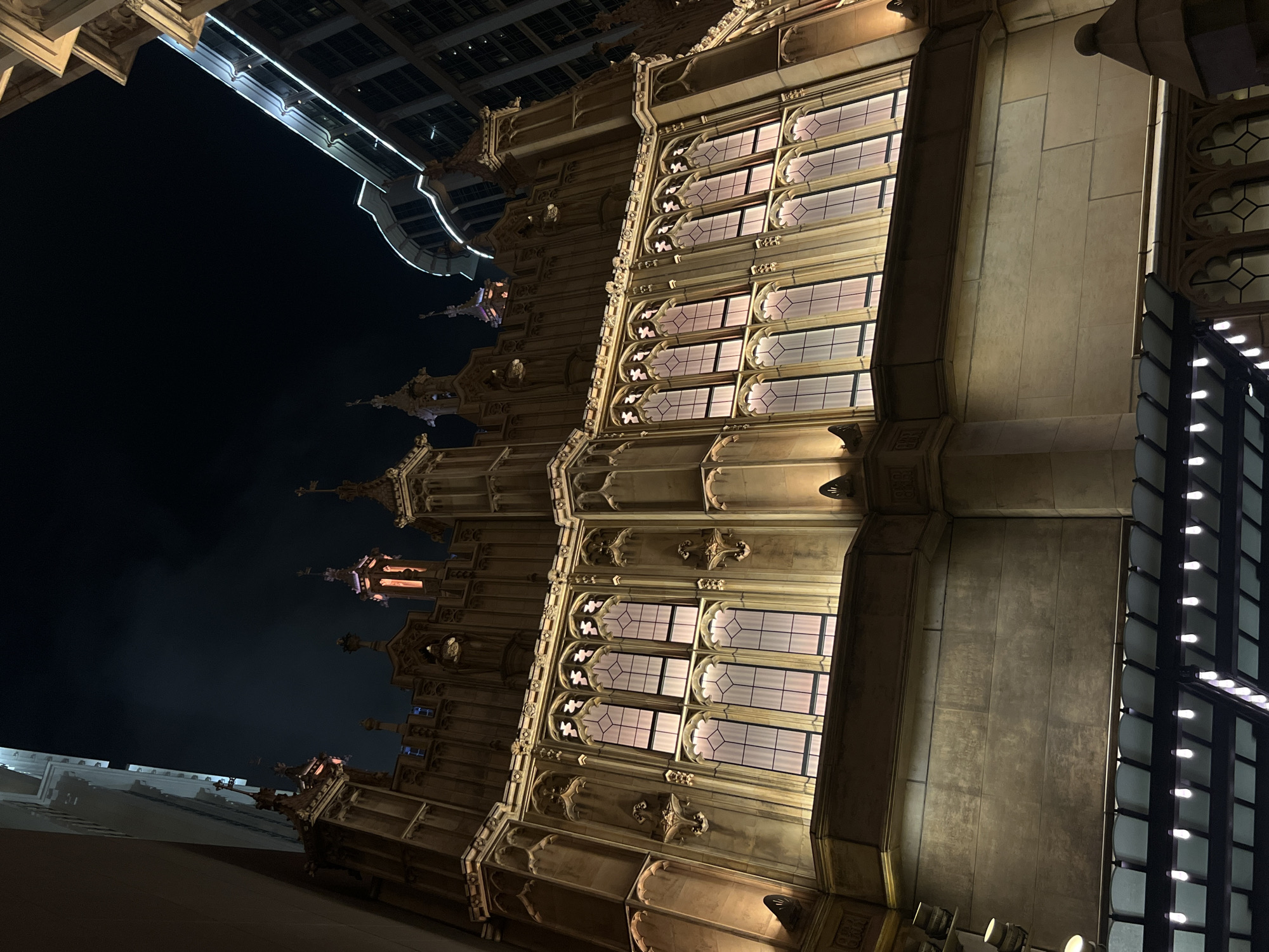 The image shows a low-angle, nighttime view of a building with ornate Gothic-style architecture. The facade features several rows of large windows, each subdivided into smaller panes with decorative tracery. Light emanates from within the building, illuminating the windows and parts of the exterior. To the left, a darker, modern building structure is visible against the night sky. The perspective is unusual, suggesting the photo was taken from the ground, looking upwards. The location, a city of Unknown, Macao, suggests a blend of historical and modern architecture in this urban setting. There are no people visible in the frame.