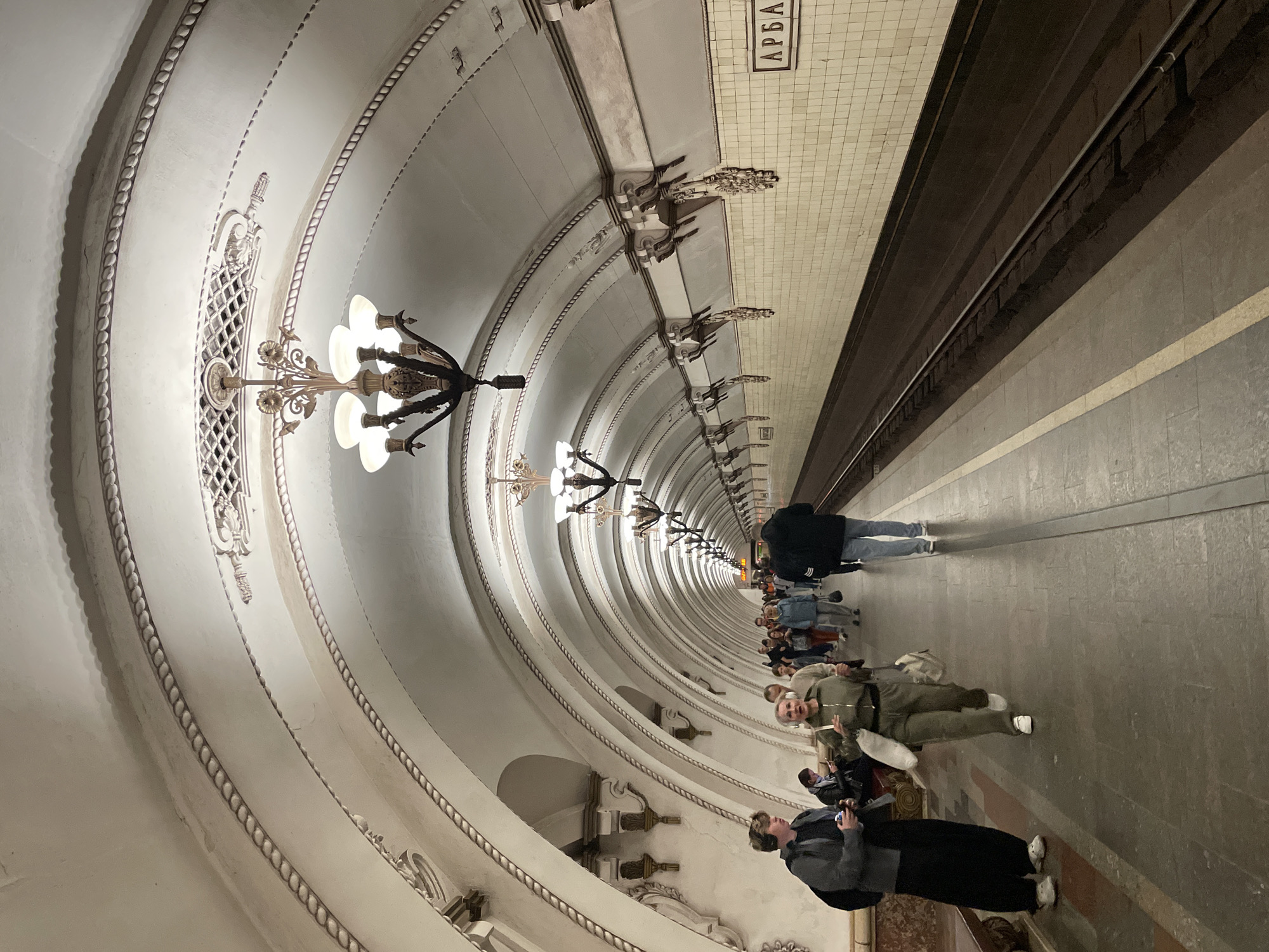 The image depicts the Arbat station on the Moscow Metro in Russia. The station has a distinctive arched ceiling with ornate chandeliers and decorative elements. People are standing on the platform, waiting for a train. The platform is wide and gray, marked with a white safety line. The walls are tiled with small squares. Above the platform, signs indicate the station name in Cyrillic: "АРБАТ".