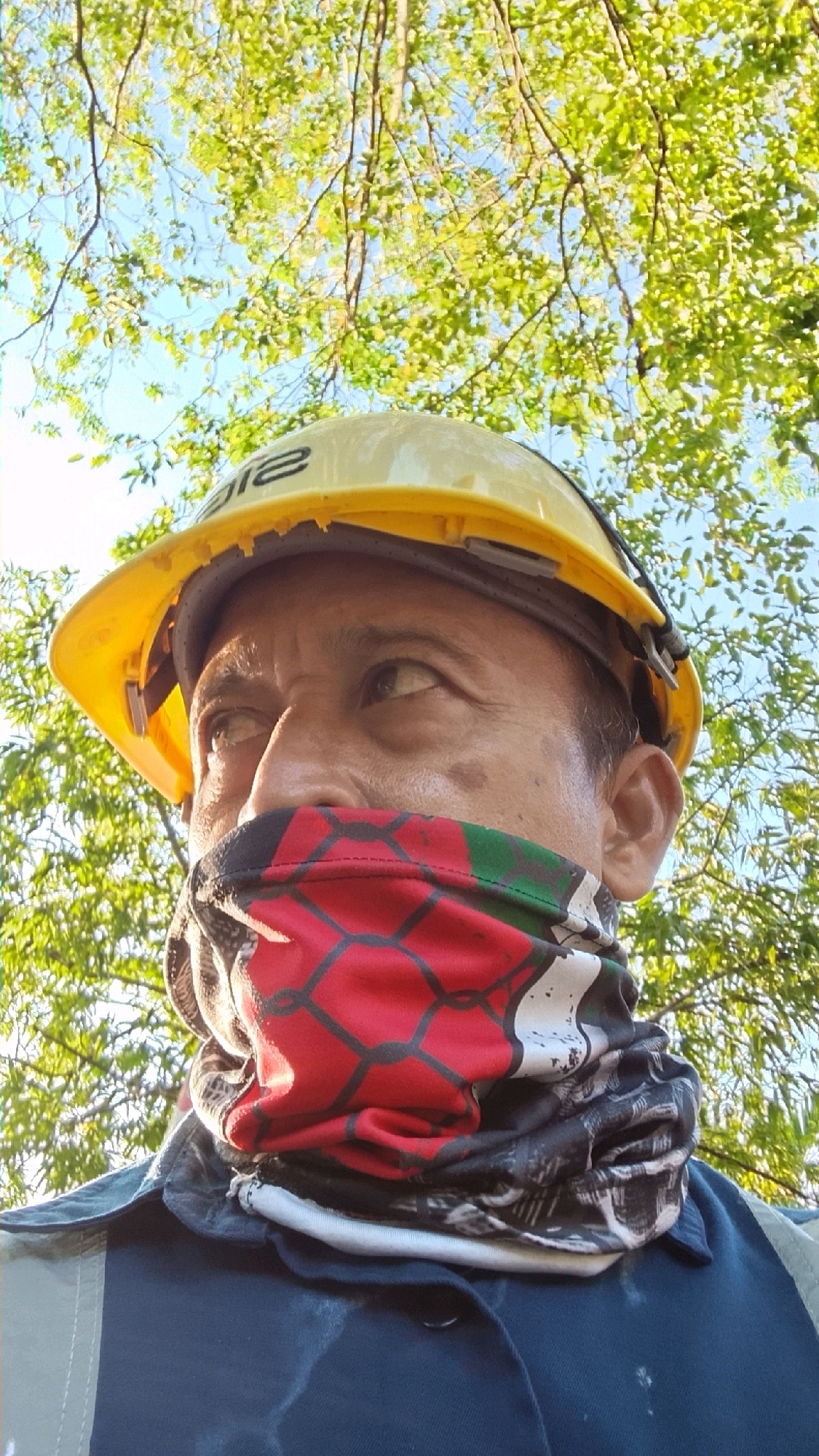 A close-up, low-angle shot shows a person wearing a yellow hard hat and a multi-colored neck gaiter covering their mouth and nose. The neck gaiter has a repeating pattern of red, black, and green sections. The person's eyes are visible above the gaiter, looking upwards. They are wearing a dark blue jacket with a light gray collar. The background consists of green foliage from trees and a bright blue sky, suggesting an outdoor setting. The image is taken in the city of Tlogobendung, Indonesia.