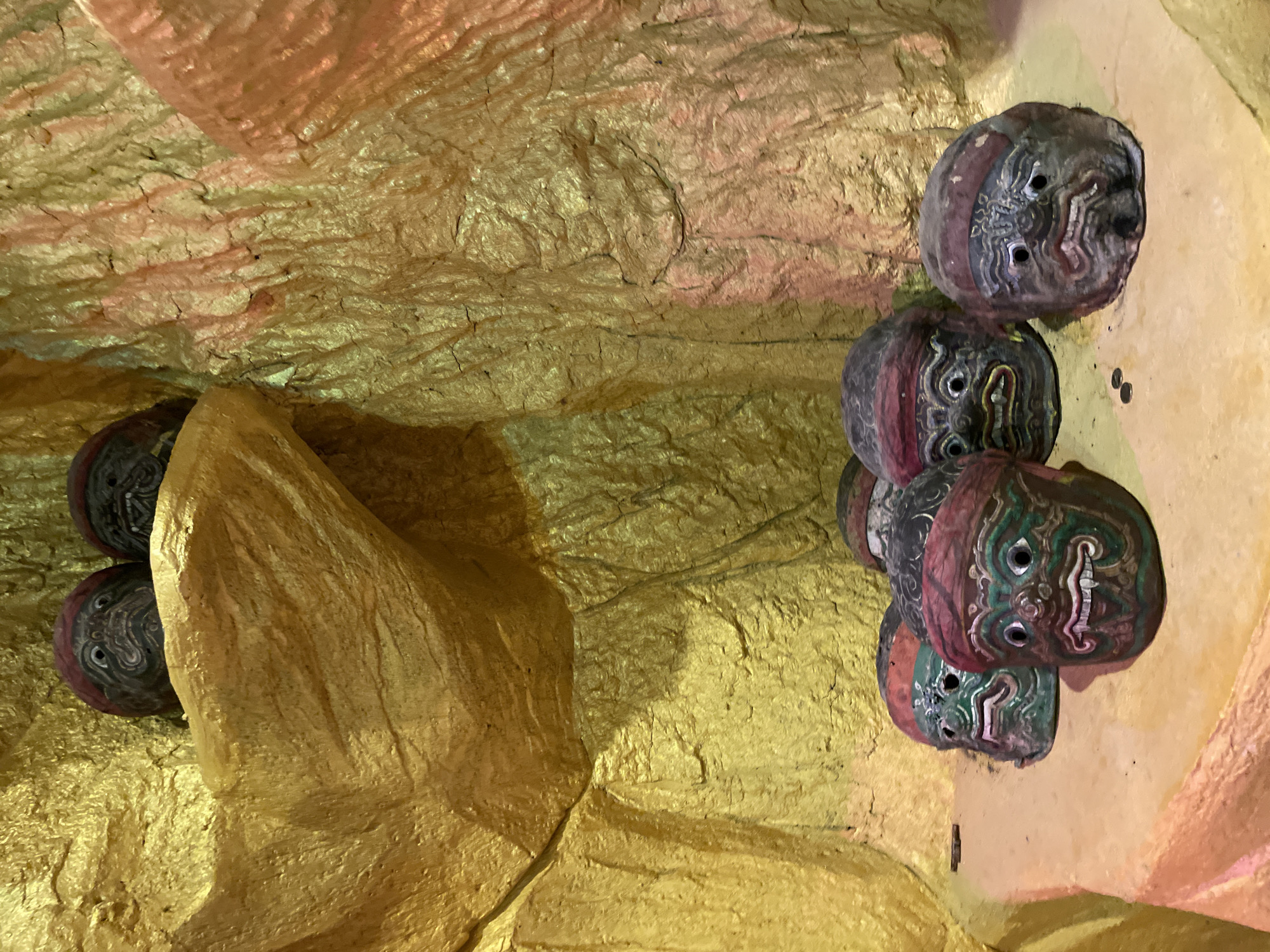 In Tambon Bo Win, Thailand, a collection of weathered masks is displayed within a cave-like structure painted in metallic gold and orange hues. Two masks are positioned high on a ledge, while five others are clustered on a sandy platform in the foreground. These masks, each with distinct facial features, painted expressions, and head coverings, appear to be made of wood or a similar material. The environment is dimly lit, with the metallic paint reflecting the light, creating a dramatic and slightly eerie atmosphere. The texture of the cave walls is rough and uneven, suggesting a natural or man-made imitation of rock.