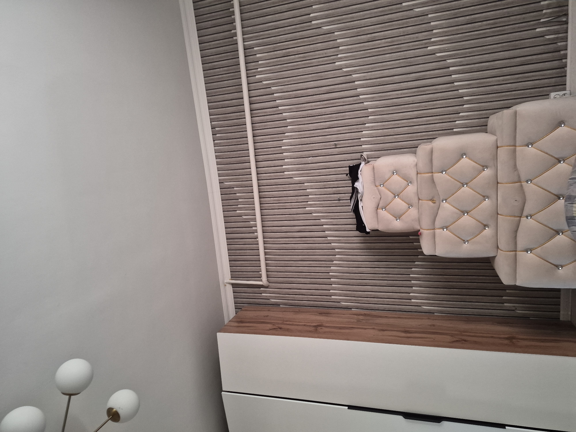 The image depicts an interior scene with a focus on a decorative wall and furniture. On the left, a section of a tall white wardrobe with a wooden accent is visible. To the right of the wardrobe, a three-tiered piece of upholstered furniture, likely a headboard or ottoman, is positioned against a wall covered in a textured wallpaper. The wallpaper features a pattern of vertical, light-colored slats with darker recessed areas, creating a rhythmic, wave-like effect. The upholstered furniture is a light beige color and features diamond-shaped tufting with gold cord trim and silver studs at the intersections. A few dark items of clothing are draped over the top tier of the furniture. In the upper left corner, a modern light fixture with multiple white globe shades is partially visible against a white ceiling. The image appears to be taken from a low angle, looking upwards. The scene is clean and organized, suggesting a residential or hotel setting. There is no indication of the city of Bereke, Kazakhstan, within the image itself.