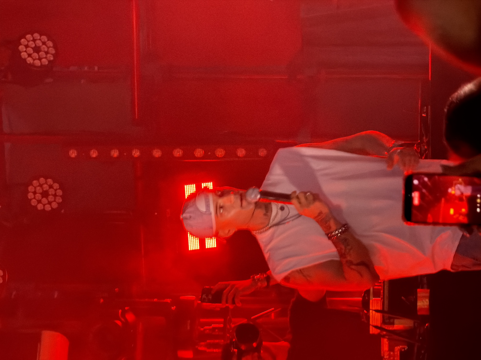 The image shows a man wearing a white sleeveless shirt and a light-colored baseball cap, holding a microphone and speaking into it. He has tattoos on his arms and neck, and is wearing necklaces. He is standing on a stage with red lighting and stage lights visible in the background. There are also people in the foreground holding up phones to record. The scene appears to be a concert or live performance. The image is stated to be from Sofia, Bulgaria.