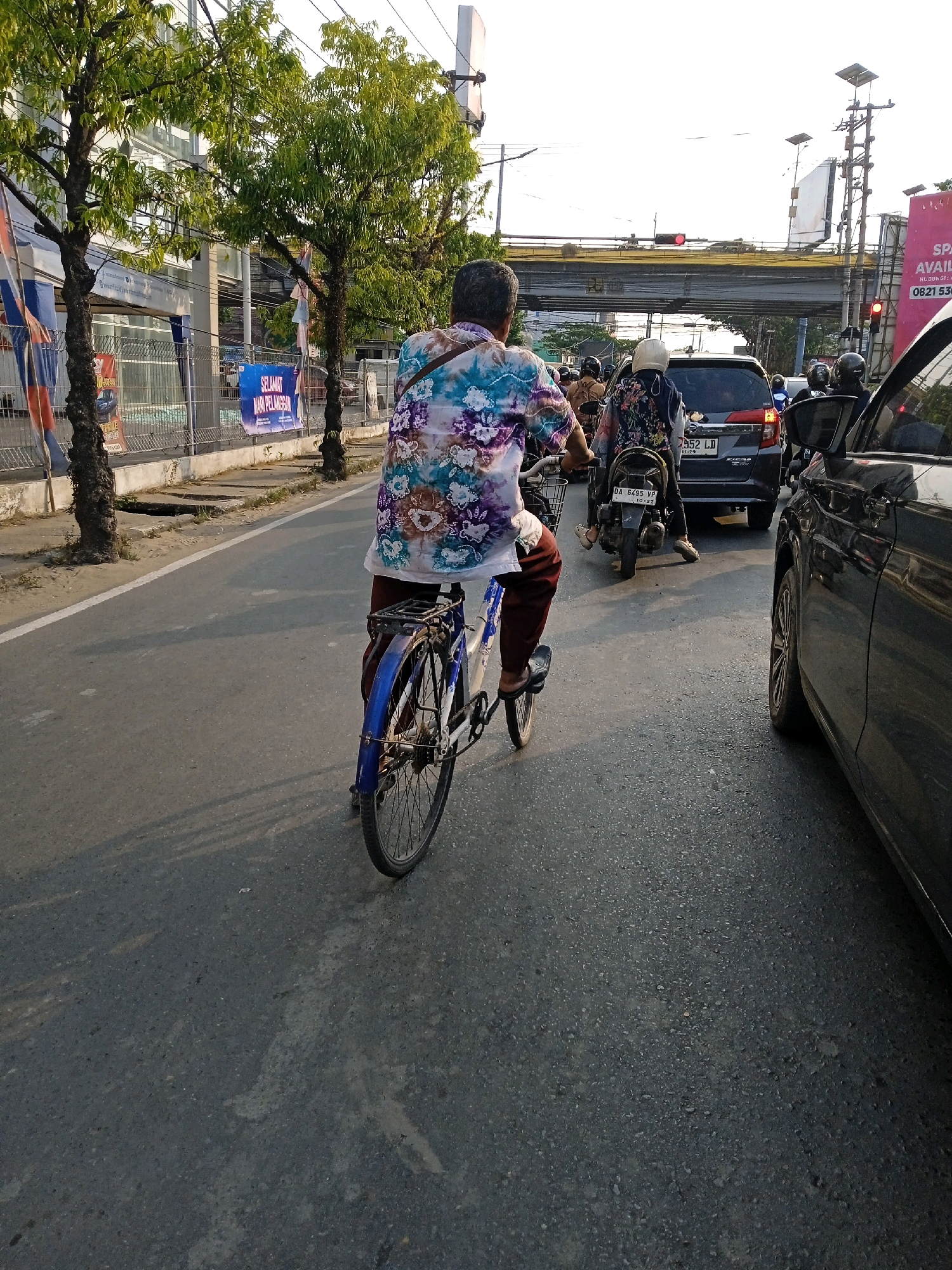 In Banjarmasin, Indonesia, on a sunny day with light traffic, a person rides a bicycle along a paved road, heading away from the camera. The cyclist, wearing a colorful floral shirt and maroon pants, is positioned in the center of the frame. To the right, a black car occupies a lane, and further ahead, a dark gray car and a motorcycle with a rider wearing a white helmet are visible in the traffic flow. The background reveals a street lined with trees, buildings, and overhead power lines. A traffic light shows red, indicating a stop. Banners, one with the text "SELAMAT HARI PELANGGAN," are displayed on the left side of the road, near a fenced area.