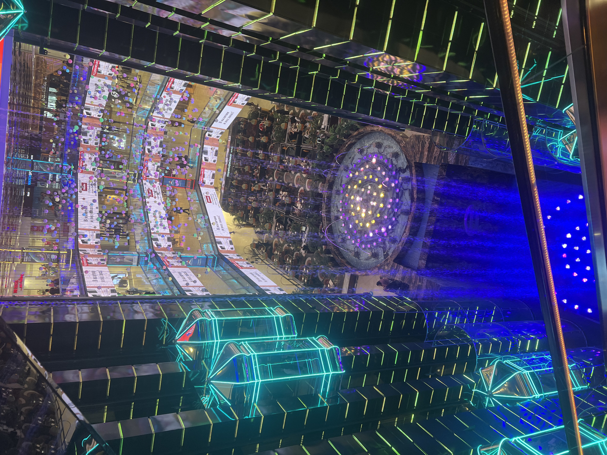 The image shows an interior space, possibly within a shopping mall or entertainment complex in Moscow, Russia. The space features reflective surfaces creating an illusion of depth and mirroring. The environment is characterized by modern, geometric designs with neon lighting in blue and green, illuminating the interior. A circular fountain with a pattern of purple and yellow lights is in the background, its water illuminated in blue. The top of the frame shows reflections of mall interiors, including shops, advertising displays, and what appear to be decorative elements like colorful balls hanging from the ceiling. The overall atmosphere suggests a technologically advanced and luxurious shopping or entertainment venue.