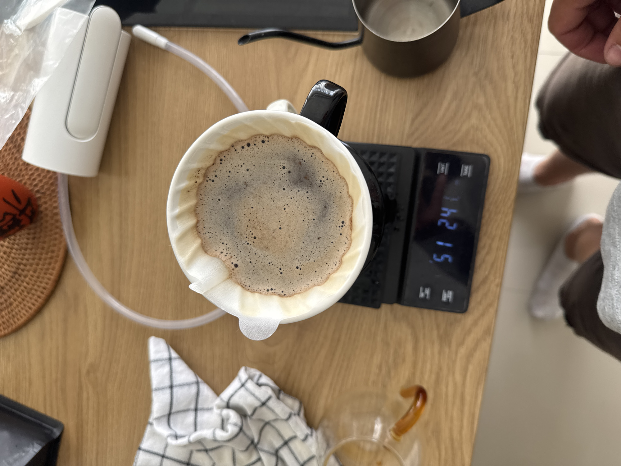 An overhead, slightly angled shot shows a close-up of a pour-over coffee setup on a wooden table. A white ceramic dripper, lined with a paper filter and filled with brewing coffee, sits atop a black digital scale displaying "51.24". To the left of the dripper, a small glass carafe with a curved handle contains a small amount of amber liquid. A black and white checkered cloth is folded to the left of the carafe. In the upper right, a white automatic pour-over kettle with a clear plastic hose is visible. A part of a person's legs, wearing white socks and brown shorts, is in the lower portion of the frame. The background shows a woven mat and other kitchen items, suggesting a domestic setting. There is no indication of the location in Kuta Selatan, Indonesia.