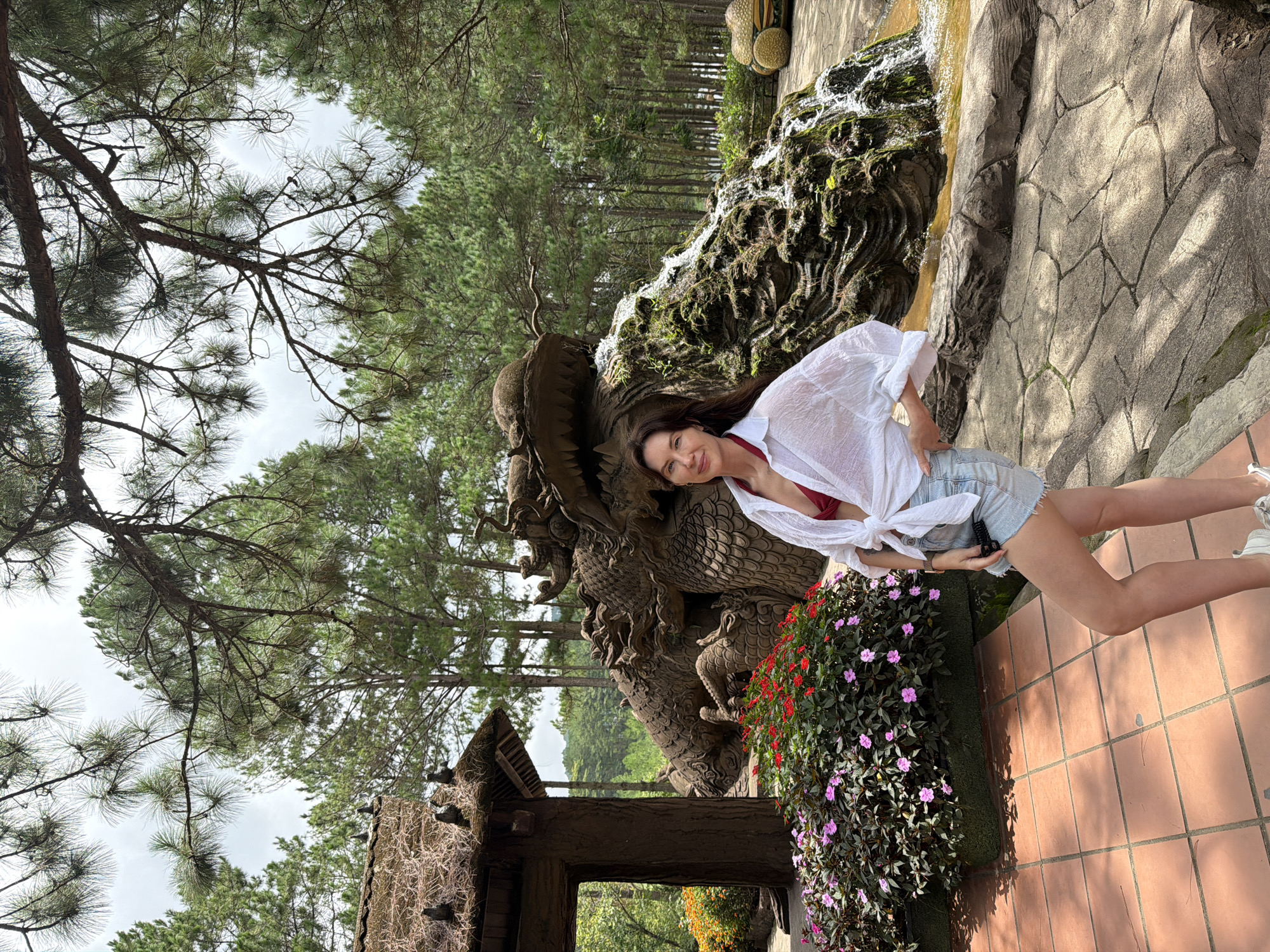 A woman stands in front of a dragon statue with a waterfall at the Dinh Thuong Lam Buddhist Temple in Da Lat, Vietnam. The woman is wearing a white unbuttoned shirt over a red bikini top, denim shorts, and white sneakers. She has long dark hair and is looking at the camera. The dragon statue is large and detailed, with moss growing on it, and the waterfall flows down its body. Behind the statue are pine trees. In the foreground, there are red and pink flowers and a tiled floor. The overall scene is outdoors on a cloudy day.