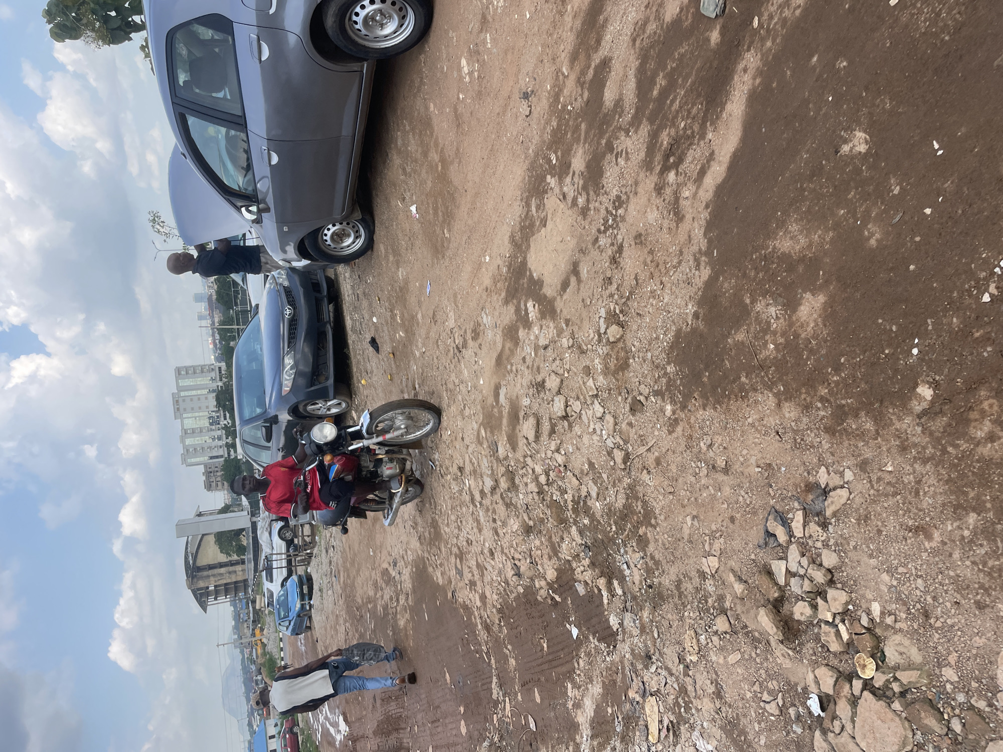The image depicts a scene in Abuja, Nigeria, characterized by a dirt road filled with potholes and debris. In the foreground, a man wearing a red shirt and black pants rides a motorcycle. To his left, another man in a white shirt and blue jeans walks away from the camera, carrying a dark object. In the background, several vehicles are visible, including a gray sedan with its hood open, suggesting mechanical work. A man in a black shirt stands near the open hood. A dark-colored car is parked to the left of the gray sedan. Beyond the vehicles, city buildings and a distant mountainous landscape are discernible under a cloudy sky. The environment appears to be an undeveloped or undergoing development area within the city.