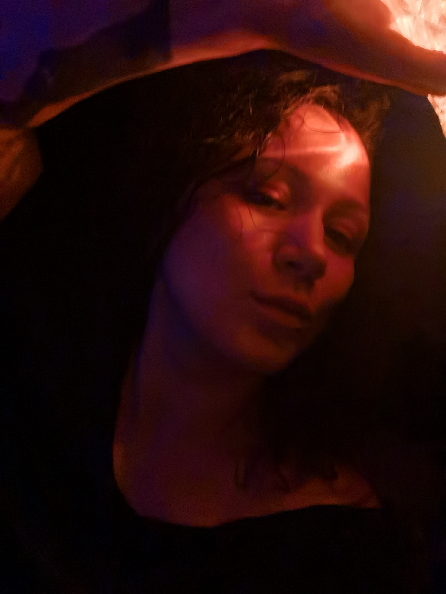 A close-up photo shows a woman with short, curly, dark hair, illuminated by a red light source. Her face is angled slightly towards the camera, and her expression appears relaxed. She has a tattoo on her chest. The background is mostly dark, with a light source on the upper right. Part of an arm appears on the top.