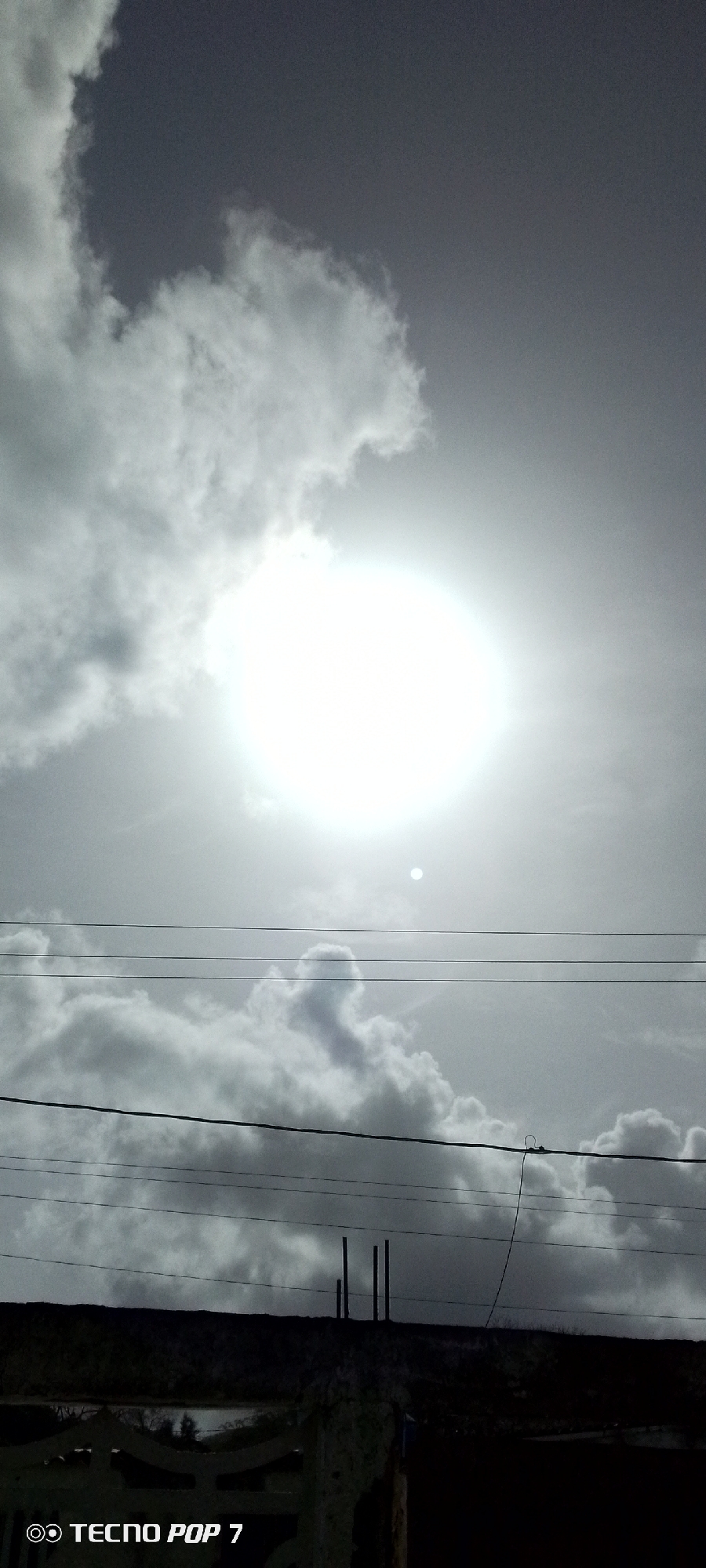 The image, possibly taken in Altagracia, Venezuela, presents a scene dominated by a bright sun partially obscured by clouds. A power line is seen in the mid-ground. In the foreground are a fence and some poles. The photo was taken with a TECNO POP 7 mobile phone.