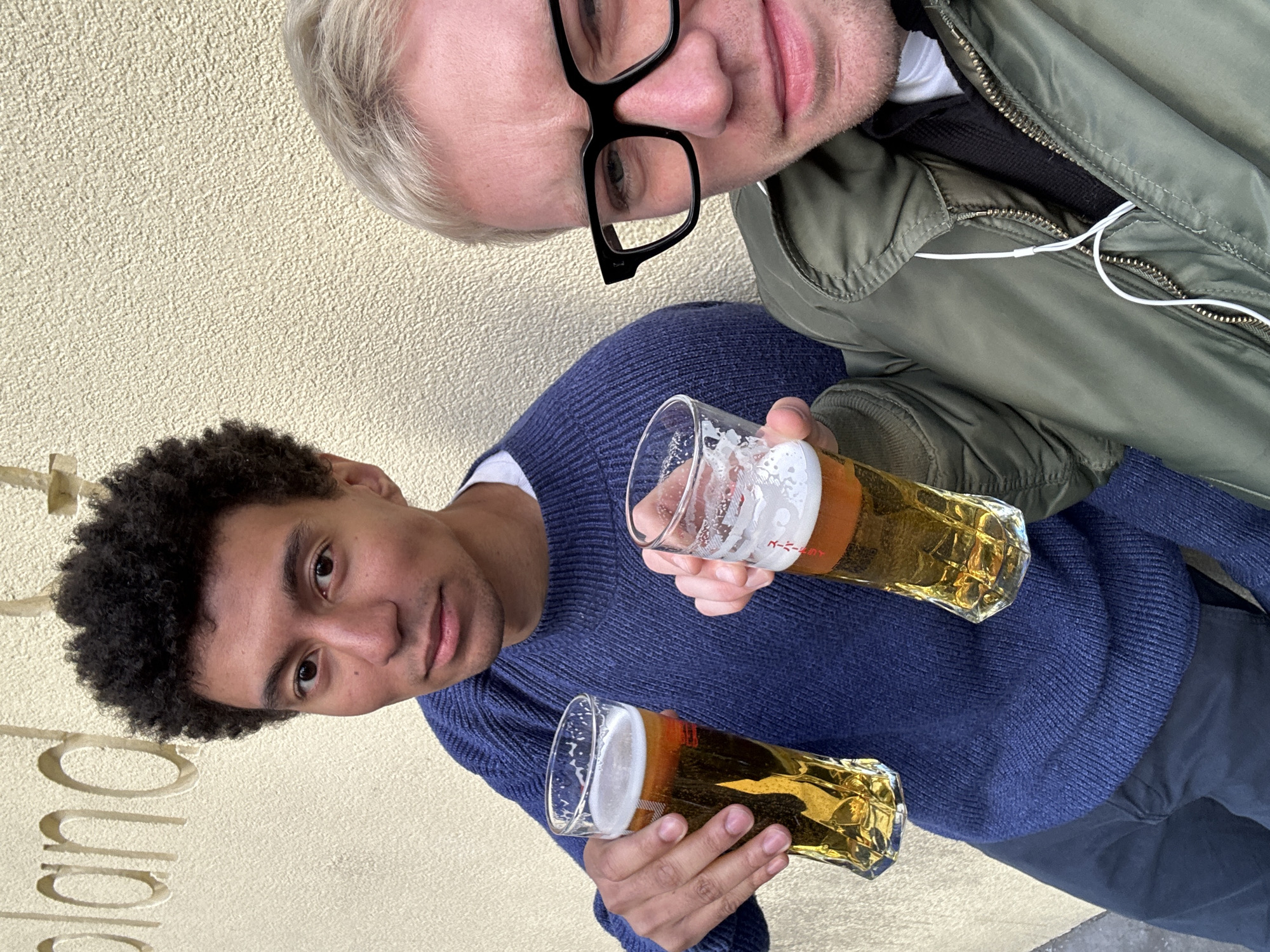 Two men pose for a selfie outdoors, each holding a glass of beer. One man has dark curly hair and wears a blue sweater. The other man has light-colored hair, wears glasses, and an open olive green jacket with white earphones. The background appears to be a textured beige wall with visible lettering. The setting is likely in London, United Kingdom, based on the style of the beer glasses.