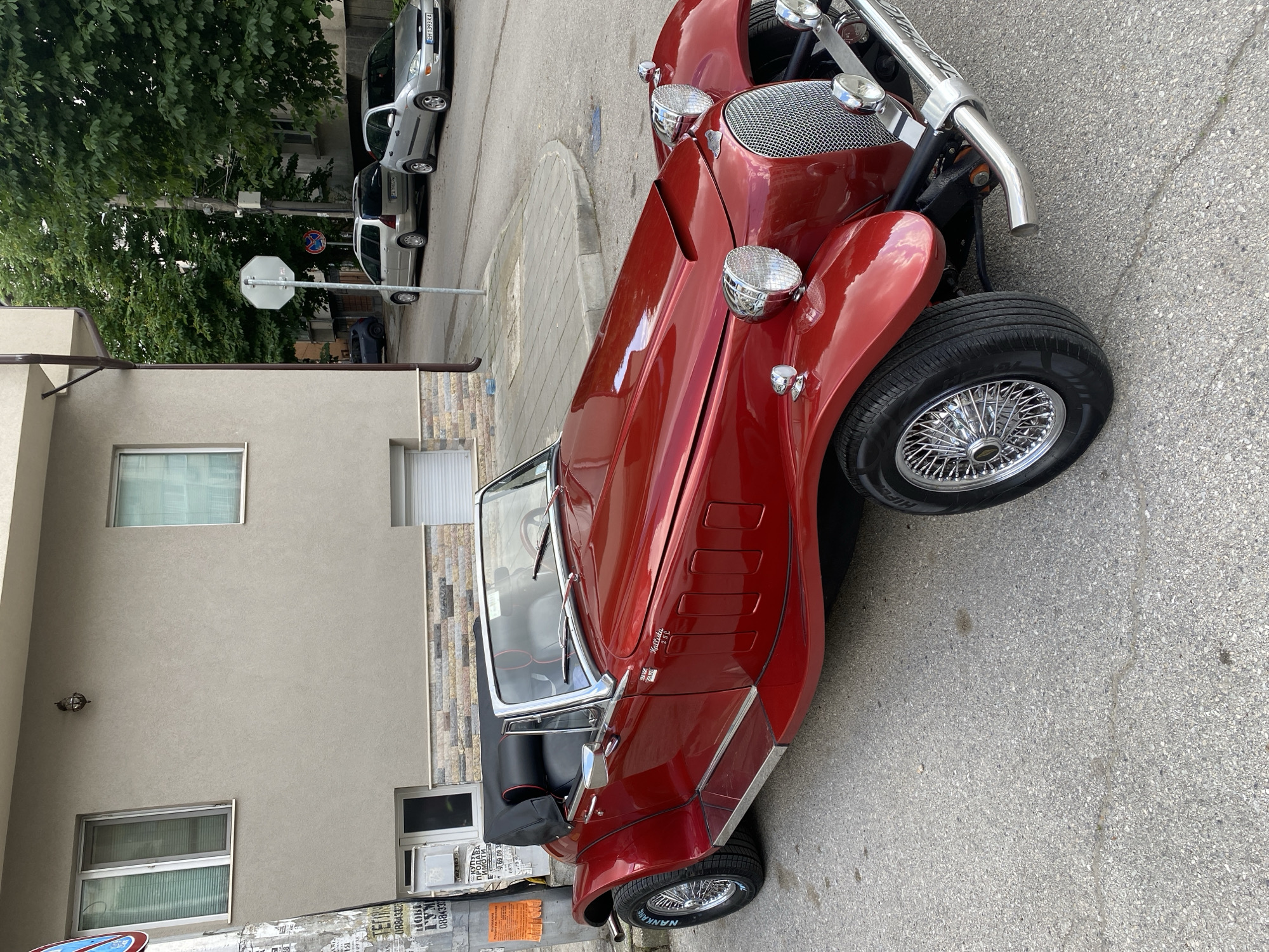 The image shows a red antique convertible car parked on a city street. The car has classic design features like wire wheels, external headlights, and chrome details. Buildings are visible in the background with posters on the walls. Cars are parked in the background. The street and sidewalk appear to be made of concrete. Some of the letters show on the tire are "NANKANG" on the front tire. The car has a sticker showing "Kallista" and "252." Based on the number plates on the cars in the background, this appears to be in Pleven, Bulgaria.