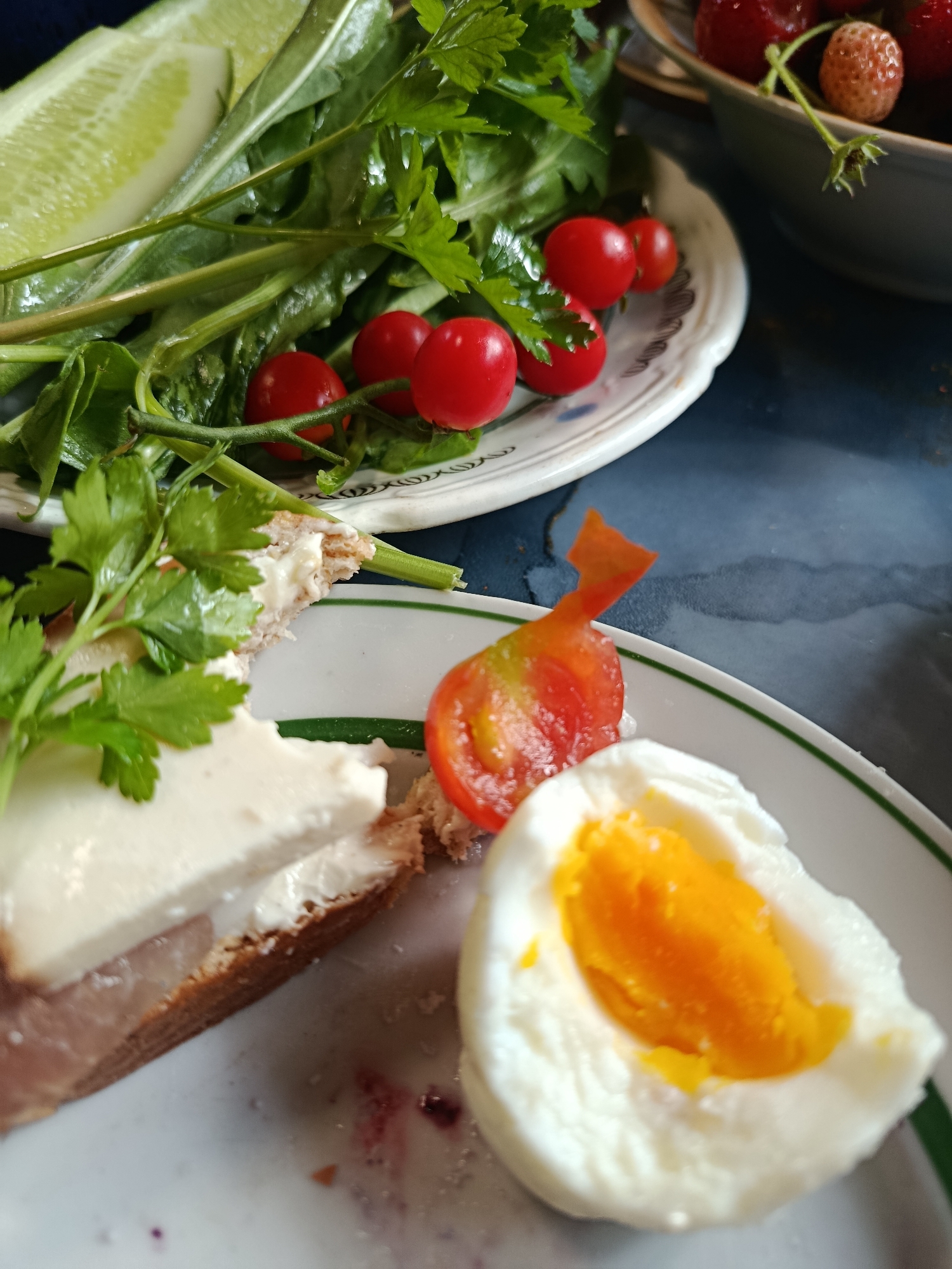 A close-up, overhead shot shows a plate of food, including a sandwich with white cheese and ham, a hard-boiled egg cut in half to reveal its yolk, and a garnish of parsley. Nearby is another plate with cherry tomatoes and a slice of cucumber with leafy greens. In the background, a bowl of strawberries is partially visible. The scene appears to be set on a dark, possibly wooden surface. The food is arranged artfully, suggesting it is a meal, possibly breakfast or lunch. The lighting is natural, casting soft shadows. There are no people or animals visible in the frame, and no indication of the specific location within Vereya, Russia.