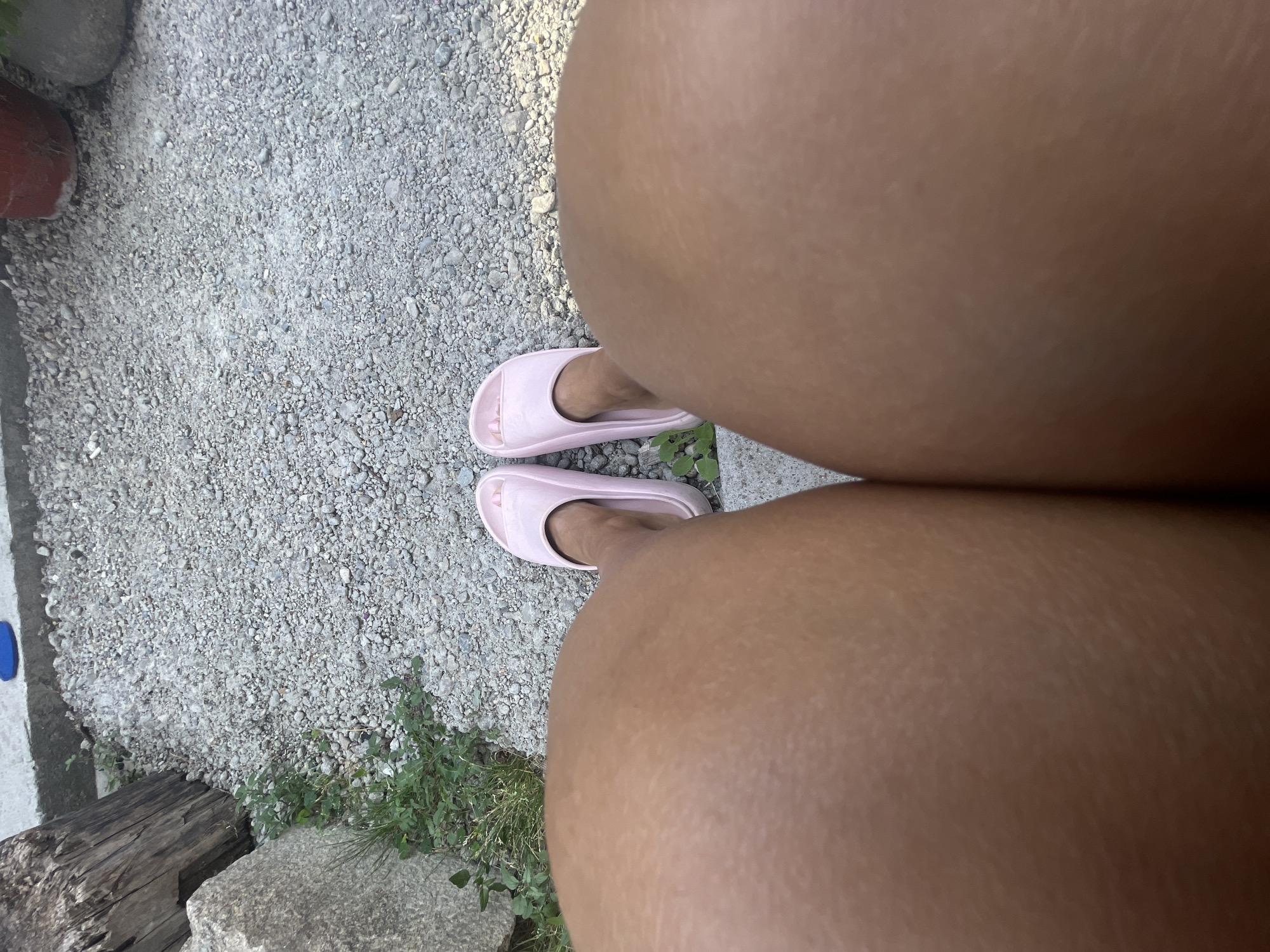 The media file shows a top-down view of a person's legs and feet wearing pink slides. The person is standing on a gravel path. To the left, a red planter and some rocks are visible. To the right, there is greenery and a concrete border. In the foreground, a piece of weathered wood and a large grey stone are partially visible. The scene appears to be outdoors, possibly in a garden or patio area. The lighting suggests it is daytime. The image is a close-up, focusing on the legs and feet, and does not provide much context about the overall activity or the person's emotions. There is no visible text in the image. The location context provided is Karlovo, Bulgaria.