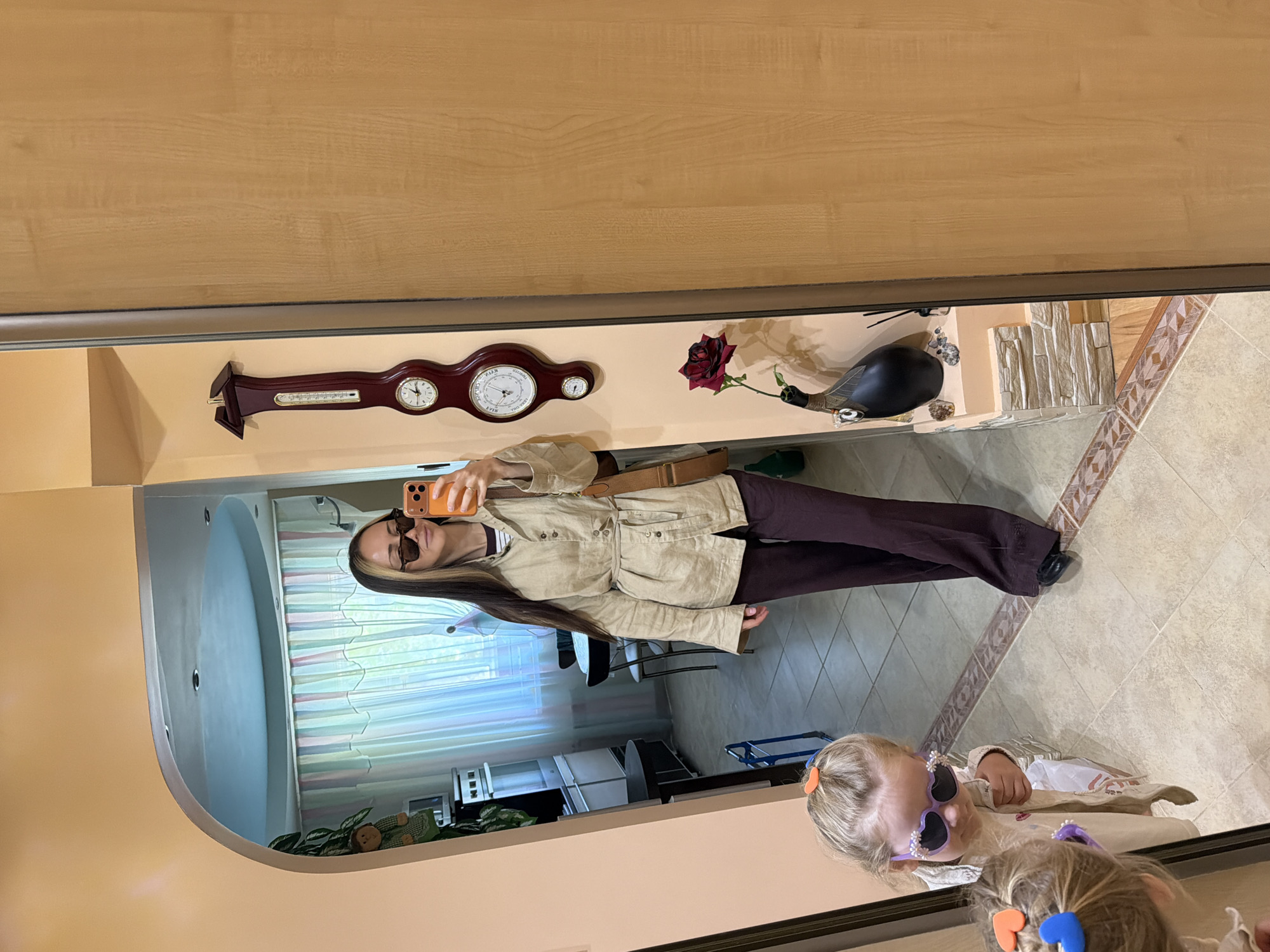 A woman with long brown hair and sunglasses is taking a selfie in a mirror, holding an orange phone. She is wearing a light brown jacket and dark brown pants. In the mirror's reflection, a young girl with blonde hair and sunglasses is lying on the floor. The background of the mirror's reflection shows a room with white and colored striped curtains and white cabinets. Above the mirror, there is a wooden weather station, a red rose, and a black decorative bird. The location appears to be an interior setting, possibly an apartment. There's no definitive evidence to confirm the location in Kamyshin, Russia.
