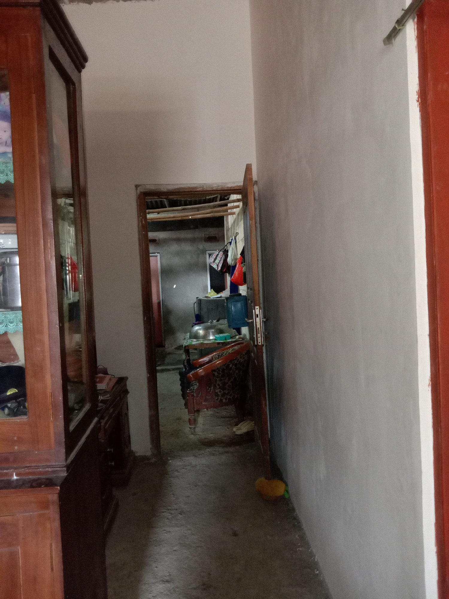 The image shows an interior hallway of a residence in Pacing, Indonesia. On the left, a tall wooden cabinet with glass doors is visible, with some items inside. To the right of the cabinet, the hallway extends into a room with an open doorway. Inside this room, a table with a large metal pot and a blue water dispenser can be seen. Various items, including bags and what appear to be kitchen utensils, are hanging on a wall in the background. The walls of the hallway are painted a light color, and the floor is concrete. A red door is partially visible on the far right side of the image.