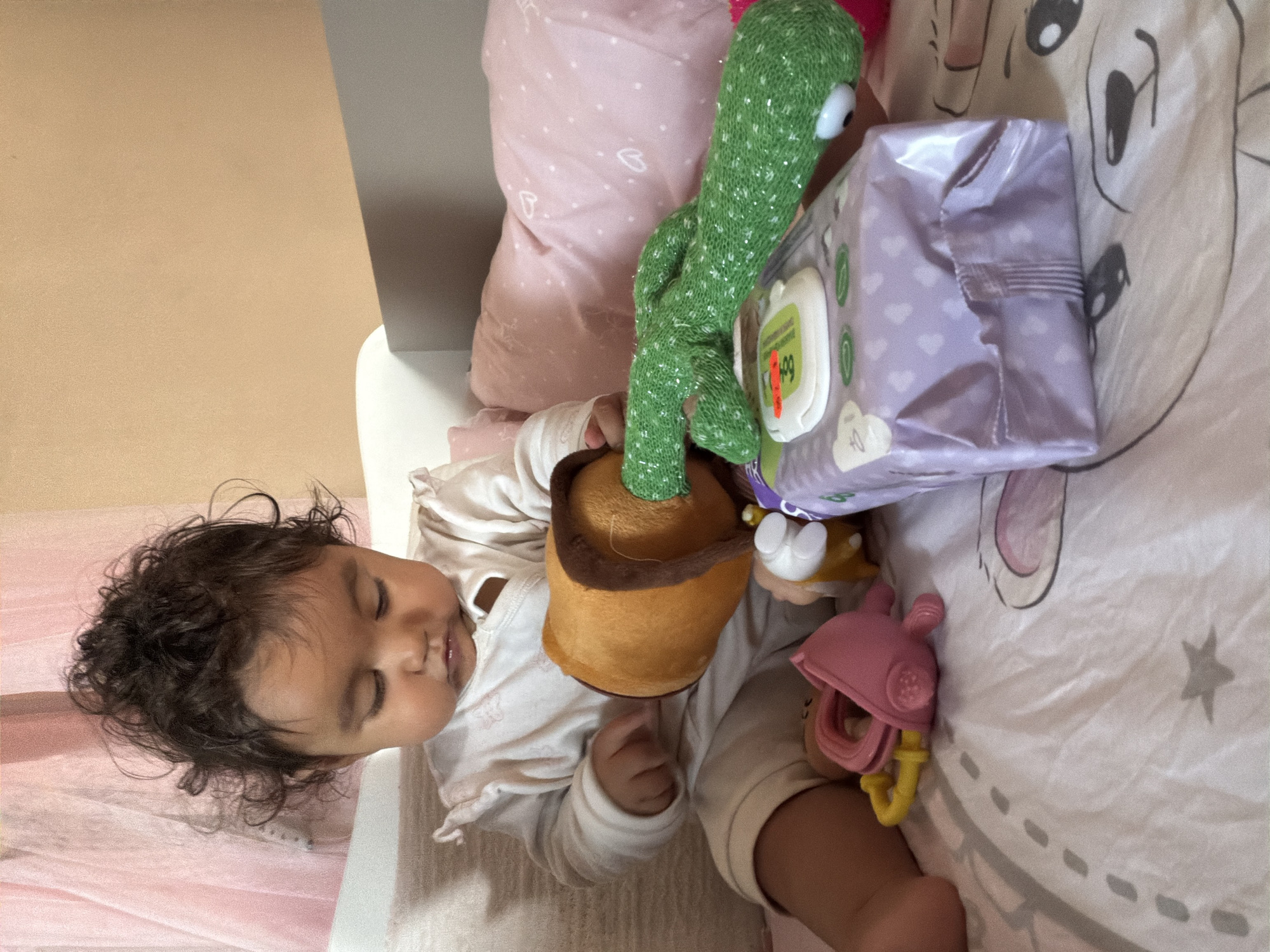 A baby is sitting on a bed covered with a white sheet depicting cartoon animals. The baby, a female with dark curly hair, is wearing a white onesie and is focused on a brown plush toy shaped like a pot with a green knitted cactus sticking out. Nearby, on the bed, are various baby toys, including a pink and yellow teething toy and a package of baby wipes. The background shows a pink sheer curtain and a peach-colored wall. The scene is indoors, likely a bedroom. The image was taken in Razhevo Konare, Bulgaria.
