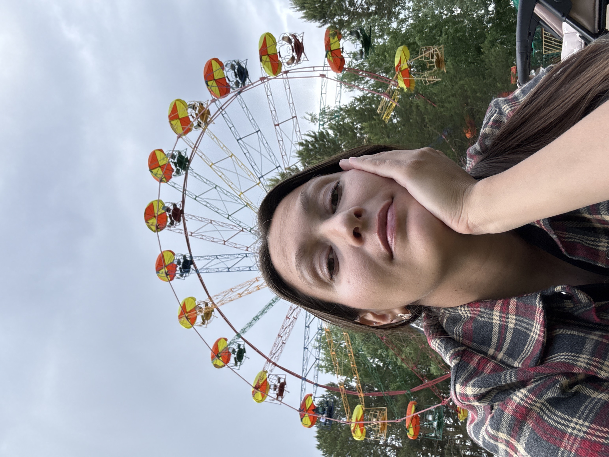 A selfie shows a woman in front of a Ferris wheel. The woman is wearing a plaid shirt and has her hand on her face. The Ferris wheel has red and yellow gondolas. Trees are visible in the background. The Ferris wheel is located in Chaikovsky, Russia.