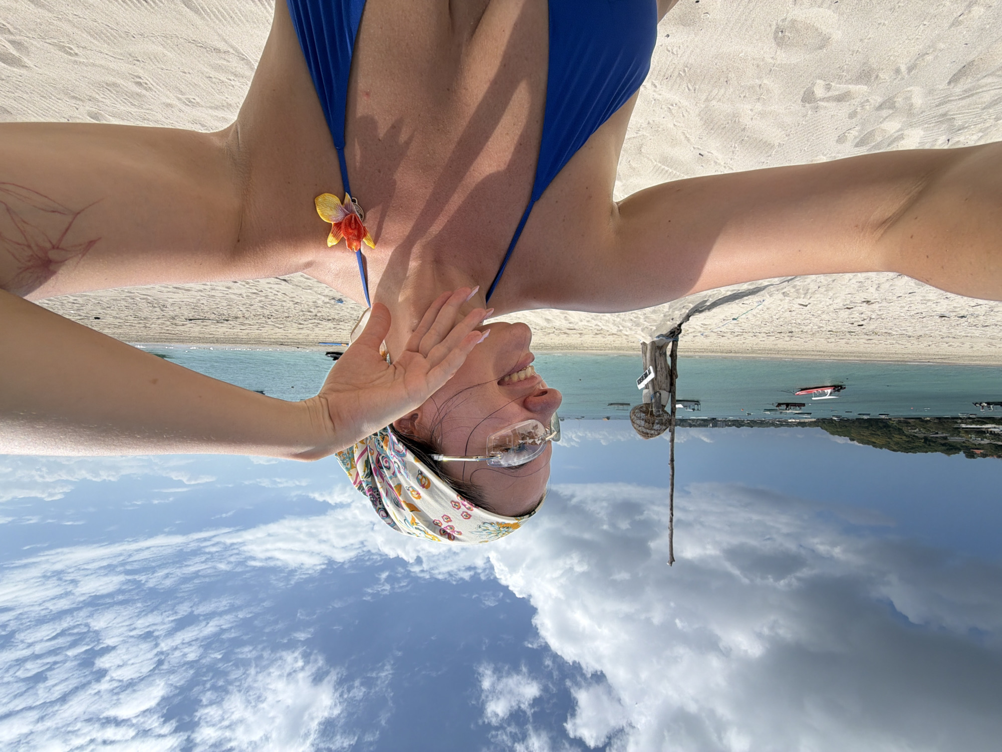 A woman, wearing a blue bikini top and a floral headscarf, takes a selfie on a sandy beach in Nusa Penida, Indonesia. She is smiling and her left arm is extended towards the camera, with her right hand resting near her neck. She is also wearing sunglasses and a flower adorns her bikini strap. The background shows a turquoise ocean with several boats, a lush green hillside with buildings, and a cloudy blue sky. A wooden post with a woven object on top is visible to the left of the woman.