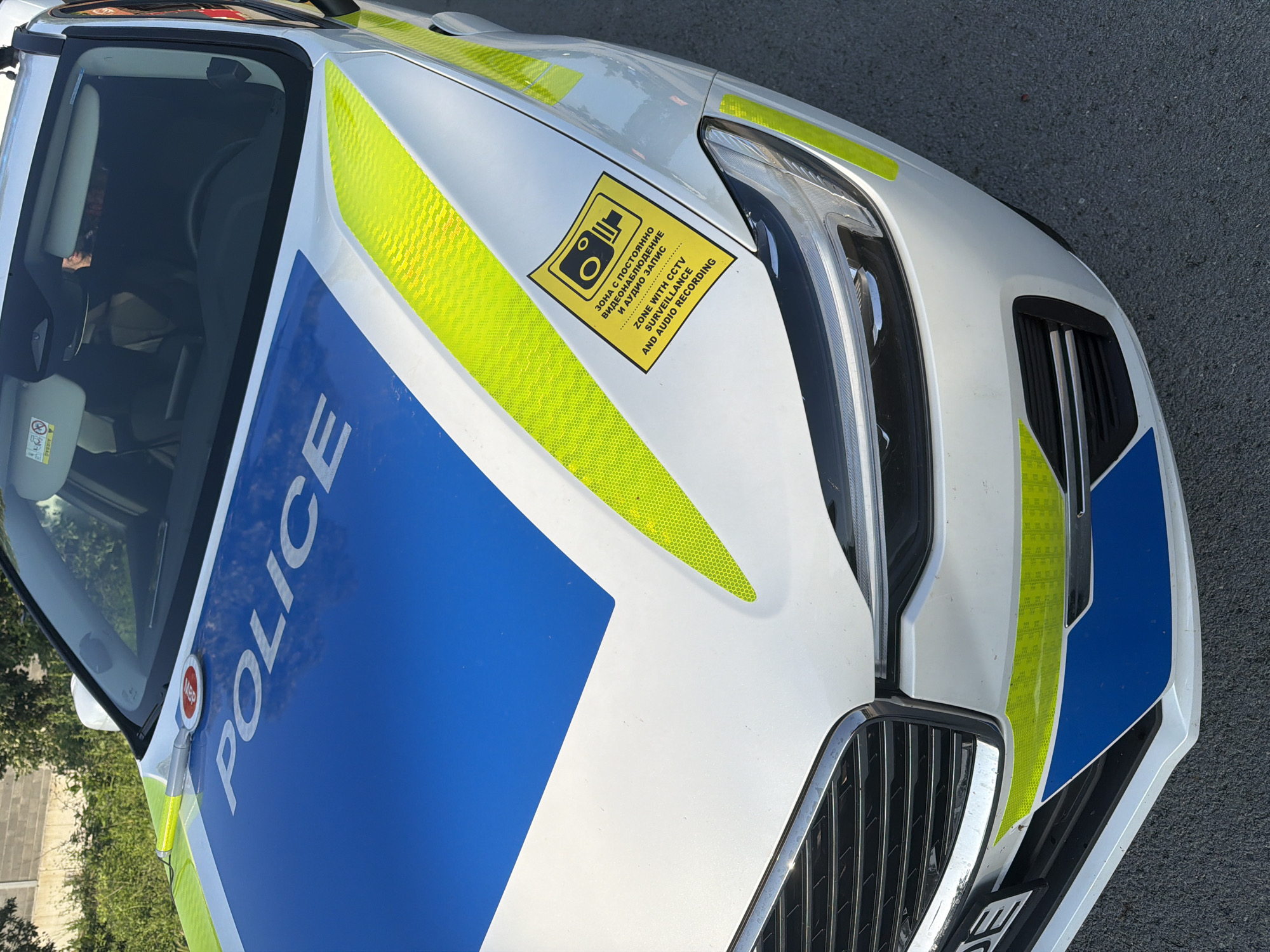 This close-up, top-down shot showcases the front-left section of a white police car, parked outdoors during what appears to be a bright, sunny day. The vehicle's hood features a prominent blue stripe with the word "POLICE" written in large white uppercase letters. Reflective yellow-green chevron patterns adorn the white areas of the hood and fender, indicating an emergency service vehicle.
A distinctive rectangular yellow sticker is affixed to the hood, near the front-left headlight. This sticker displays a camera icon and text in two languages. The Cyrillic inscription reads: "ЗОНА С ПОСТОЯННО ВИДЕОНАБЛЮДЕНИЕ И АУДИО ЗАПИС." Below it, the English translation states: "ZONE WITH CCTV SURVEILLANCE AND AUDIO RECORDING," indicating that the area around the vehicle is under constant video and audio monitoring.
The car's modern headlight assembly and part of the front grille, featuring chrome horizontal bars, are visible on the right side of the frame. Through the windshield, reflections of the sky and some internal car elements, including a small airbag warning label, can be seen. The vehicle appears stationary, with no activity taking place beyond its presence. The combination of the Cyrillic text and the police livery suggests the car is located in an Eastern European country, consistent with Shumen, Bulgaria.