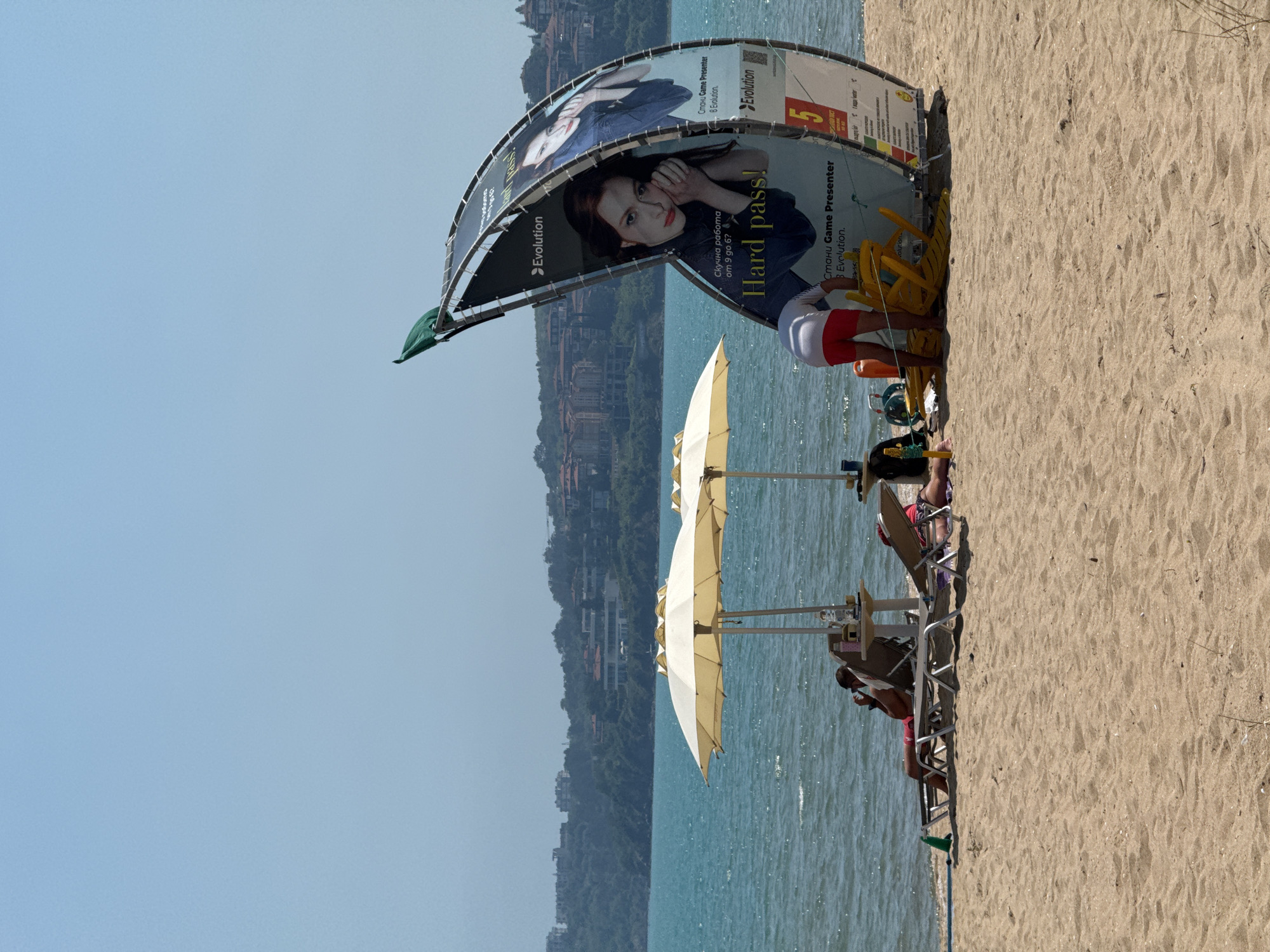 This image depicts a sunny beach scene in Sozopol, Bulgaria, featuring individuals, beach furniture, and a large advertisement by the water's edge. The foreground presents a sandy beach with subtle ripple marks and scattered small shells. On the left, a person reclines on a light-colored sun lounger, facing right, wearing red swimwear bottoms and holding a dark object to their ear. A green bottle lies beside the lounger, with a black backpack partially visible to its left. To the right, another individual, seen from the waist down, wears red shorts and a white top, bending over a stack of yellow plastic chairs and orange buoy-like objects.
In the midground, two large, light yellow or off-white beach umbrellas are open, standing upright near the water. Small, square tables are positioned beside the loungers. A prominent, tall, and curved advertising structure, resembling a half-banana or sail, stands on the sand to the right. A green flag or piece of fabric is attached to its uppermost point. The advertisement displays two female models; the left model rests her chin on her hands, looking directly forward, while the right model looks slightly upwards. Text on the advertisement includes "Evolution," "Hard pass!", and the Cyrillic phrase "Стани Game Presenter В Evolution" (Become a Game Presenter at Evolution). The water beyond the beach is a vibrant turquoise-blue, exhibiting slight surface ripples.
The background reveals a distant, hilly coastline covered in green vegetation, featuring numerous multi-story buildings with red-tiled roofs. The sky above is a pale, clear blue, transitioning to a lighter, hazy tone near the horizon, indicative of a bright, sunlit day.