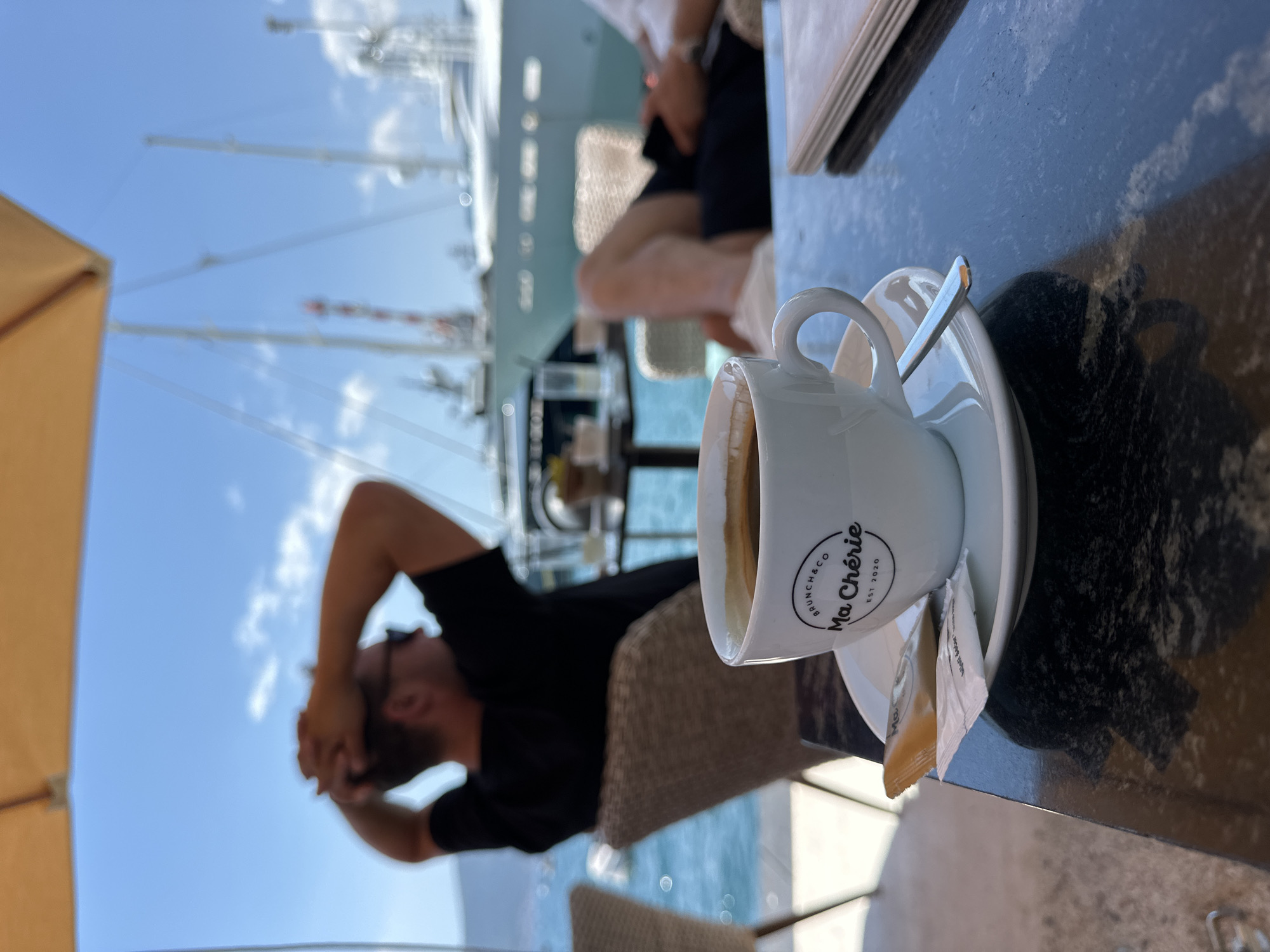A close-up shot focuses on a white cup of coffee on a black marble table. The cup has a logo that reads "BRUNCH & CO Ma Chérie EST 2020". Next to the cup are two packets, one yellow and one white, presumably sugar. A silver spoon rests on the saucer. In the background, slightly out of focus, two men are seated at tables. The man on the left is wearing a black t-shirt and sunglasses, with his hands behind his head. The man on the right is wearing a white shirt and dark shorts, looking at his phone. The setting appears to be an outdoor cafe by the water, with blue water and a large white yacht visible. The sky is blue with scattered clouds. This scene is located in Donja Lastva, Montenegro, likely at a waterfront establishment.