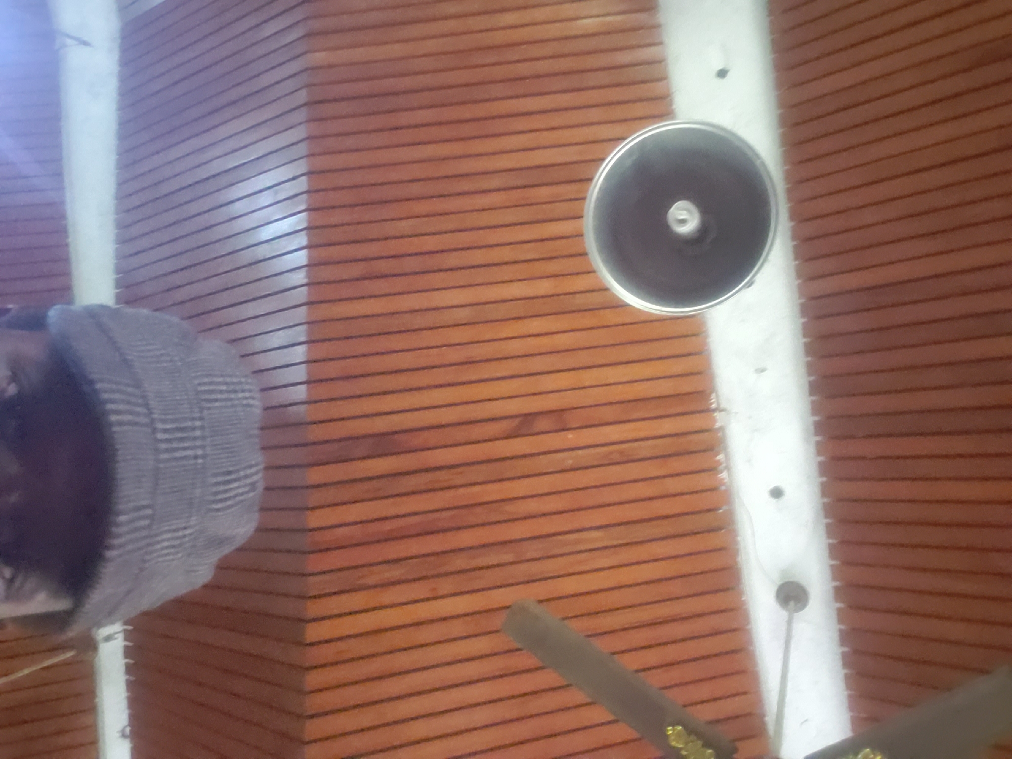 An indoor shot shows a person wearing a checkered cap, viewed from a low angle. The person's face is partially visible. Above, a ceiling fan with brown blades is partially in frame, mounted on a wooden paneled ceiling. A circular fixture, possibly a speaker or light fixture, is also visible on the ceiling. The overall setting appears to be a public space with wooden architectural elements. The location is identified as Lagos, Nigeria.