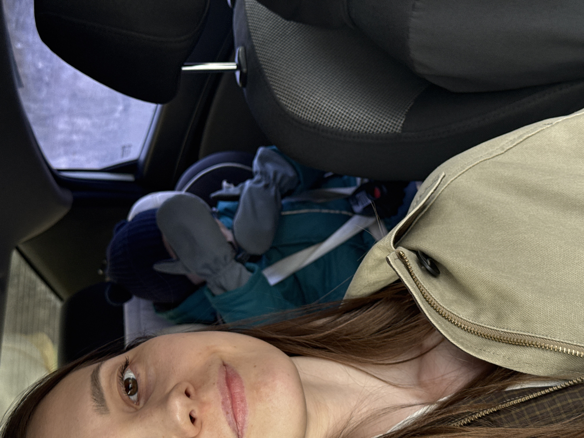 The image captures an interior view of a vehicle in the city of Chaikovsky, Russia. In the foreground, the right side of a female's face is visible, showing a brown eye, part of her nose, and her mouth. She has brown hair, a neutral expression with slightly parted lips, and is wearing a light-colored top under a brown plaid garment, overlaid by a beige or khaki jacket with a brass zipper and a dark button. In the midground, positioned in a rear passenger seat behind the front passenger's seat, a young child is seated in a car seat. The child wears a dark blue knit hat with ear-like protrusions, a teal winter jacket, and grey mittens, with their face obscured. A safety harness from the car seat is visible across the child's shoulder. The car's interior features black upholstered seats, including a headrest for the front passenger seat in the right foreground. Through the rear passenger window, a blurred, light-toned outdoor environment is visible, indicative of an overcast or snowy day. No direct interaction between the subjects is depicted.