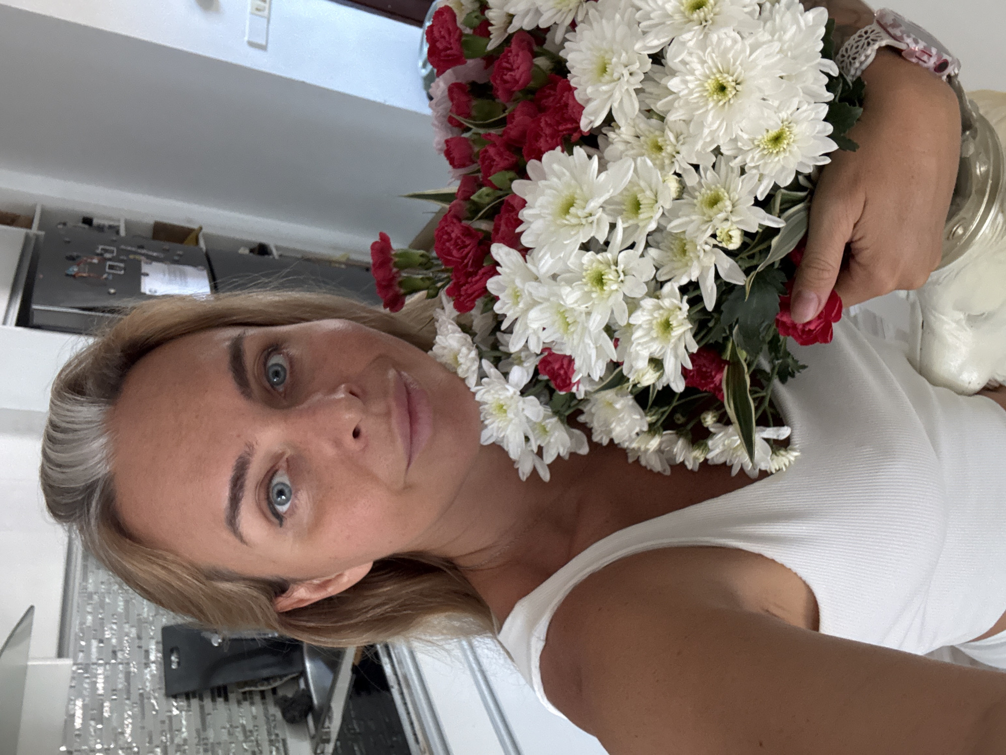 A woman stands in a kitchen, holding a bouquet of white and red flowers. She is wearing a white tank top. The kitchen has a modern design with white cabinets and a tiled backsplash. In the background, a refrigerator is visible with various items attached to its door. A sink and a stovetop are also present. The lighting suggests it is daytime.