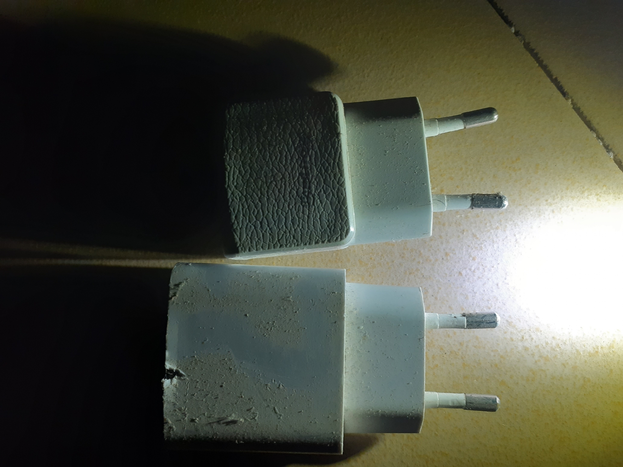 The image shows two white European-style electrical plugs against a textured, light-colored surface, possibly a wall or tabletop. The top plug appears cleaner and has a textured, dark gray surface on one side, possibly a grip or branding. The bottom plug is visibly dirty, with scuffs, dust, and what looks like a small chip or crack on its side. Both plugs have two metallic prongs extending from them. The lighting in the image creates strong shadows, with a bright light source coming from the right, causing some glare and overexposure in that area. The overall scene is domestic, depicting common electronic accessories. There is no indication of the city of Jalingo, Nigeria in the image.