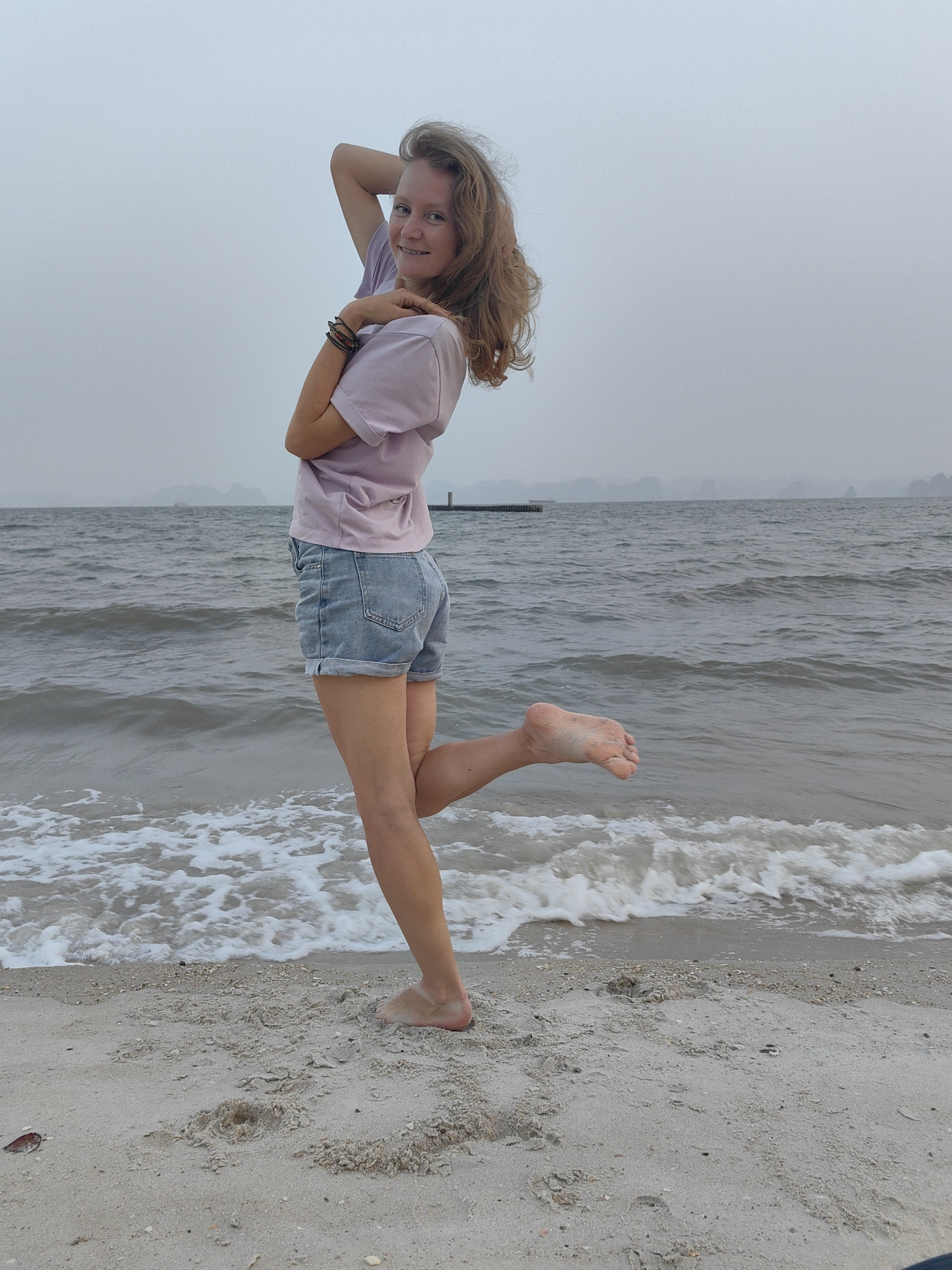 A woman with light skin and reddish-blonde hair stands on a sandy beach, partially submerged in the shallows of the sea. She wears a lilac T-shirt and denim shorts. Her left leg is raised, foot hovering above the water. Her right arm is raised above her head, while her left arm is bent across her chest. The background features a gray, overcast sky and a body of water with small, rocky islands in the distance. A structure resembling a dock is visible in the water behind her. The sand is a light gray color. The scene appears to be near Bai Chay Ward, Vietnam, based on the location.