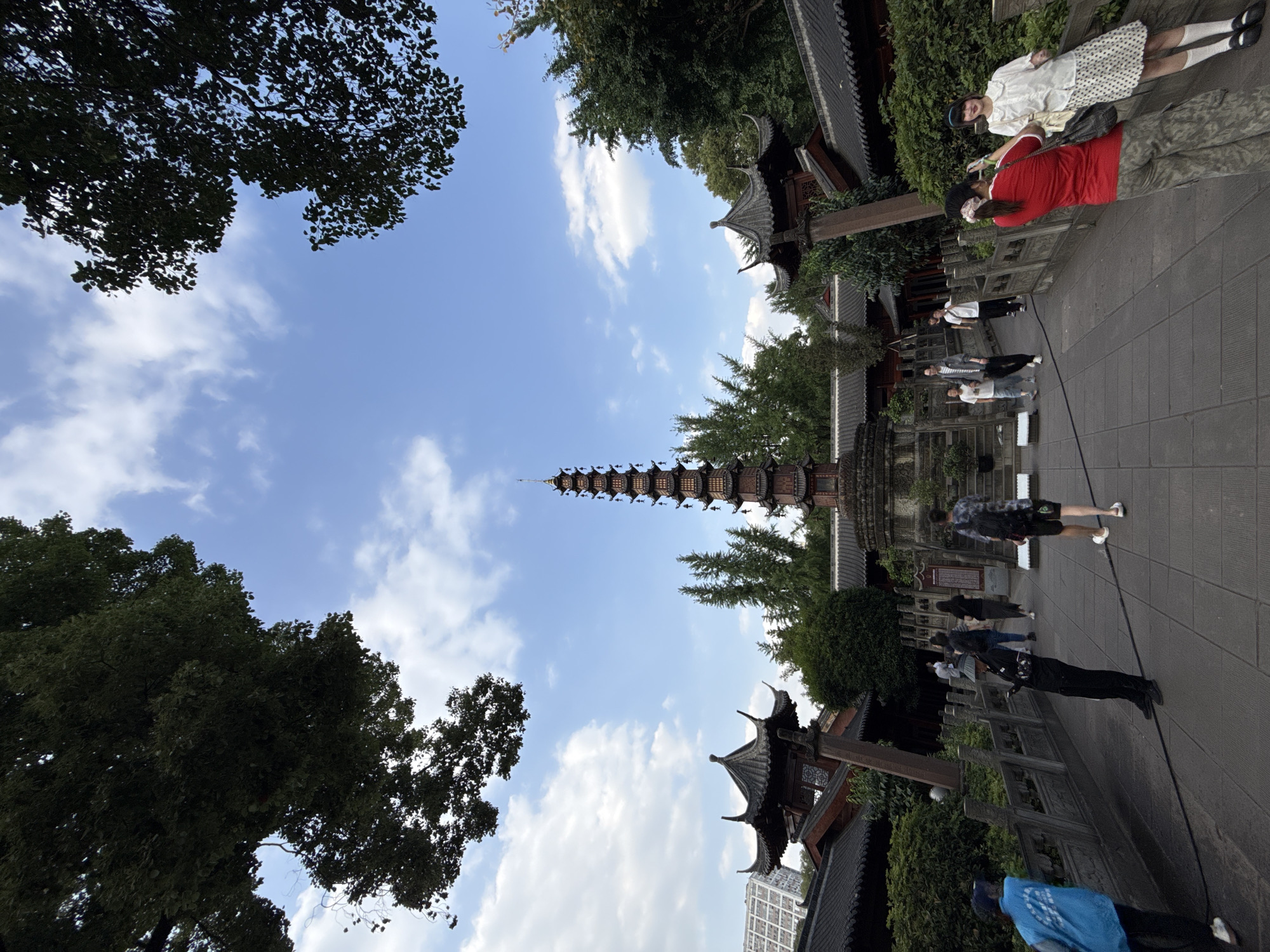 The image shows a vertical shot of a cityscape in Qingyang District, China. The sky is a clear blue with scattered white clouds. A tall, multi-tiered pagoda is centrally positioned, reaching towards the sky. The pagoda is surrounded by trees and traditional Chinese architecture with dark tiled roofs. People are walking along a stone pathway adjacent to the buildings. Some are standing and appear to be interacting. The bottom left and top left corners feature trees with dense foliage, framing the shot. A building of modern architecture is visible at the bottom right.