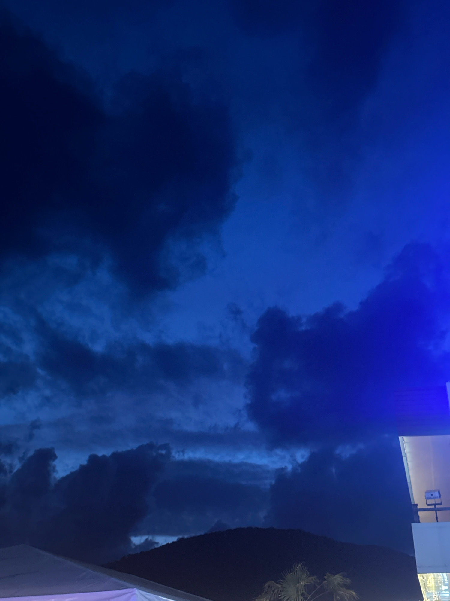This is a vertical, outdoor, low-angle shot taken at dusk or night in Lermontovo, Russia. The scene is dominated by a dark blue, cloudy sky. In the foreground, the edge of a white tent structure is visible in the bottom left corner. To the right and center of the image, a dark silhouette of a mountain range is present. A bright blue light emanates from the top right corner of the frame, illuminating a portion of a white building. A palm tree is visible in the bottom right, silhouetted against the dark sky. The overall atmosphere is dark and dramatic, with the blue light creating a stark contrast against the natural darkness.