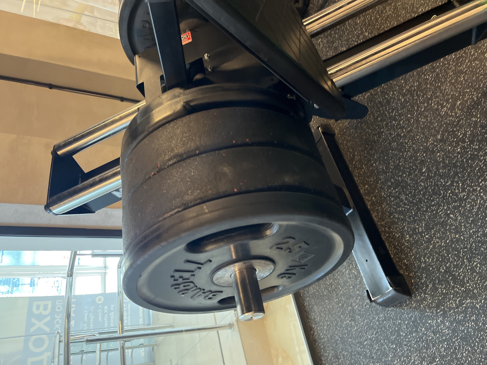 The image shows a close-up of a leg press machine in a gym. The machine has several heavy weight plates stacked on its bar. The weight plates are black and circular, with the word "BARBELL" and weights in kilograms embossed on them. The floor of the gym is black and textured. In the background, through a glass door, there is a sign with Cyrillic text, indicating the location is likely Russia. The lighting in the gym appears to be artificial, casting a warm glow on the walls. There are no people visible in the image.