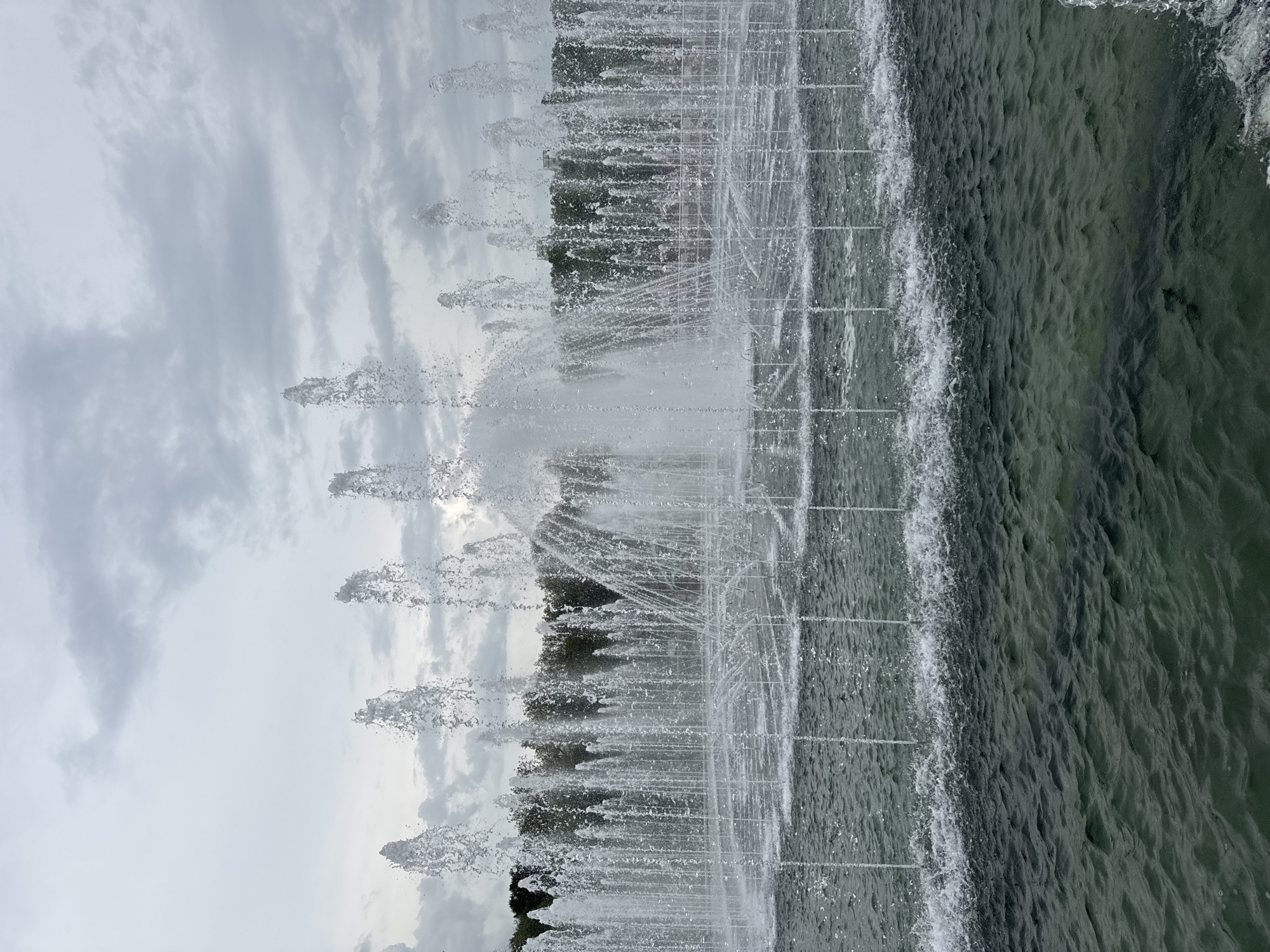 The image shows a fountain in Moscow, Russia, with multiple jets of water spraying upwards. The water creates a dense wall of spray, partially obscuring trees and the sky in the background. The sky is overcast with gray clouds. The water in the pool appears dark and rippled. The photograph is taken at a low angle, looking up at the fountain.