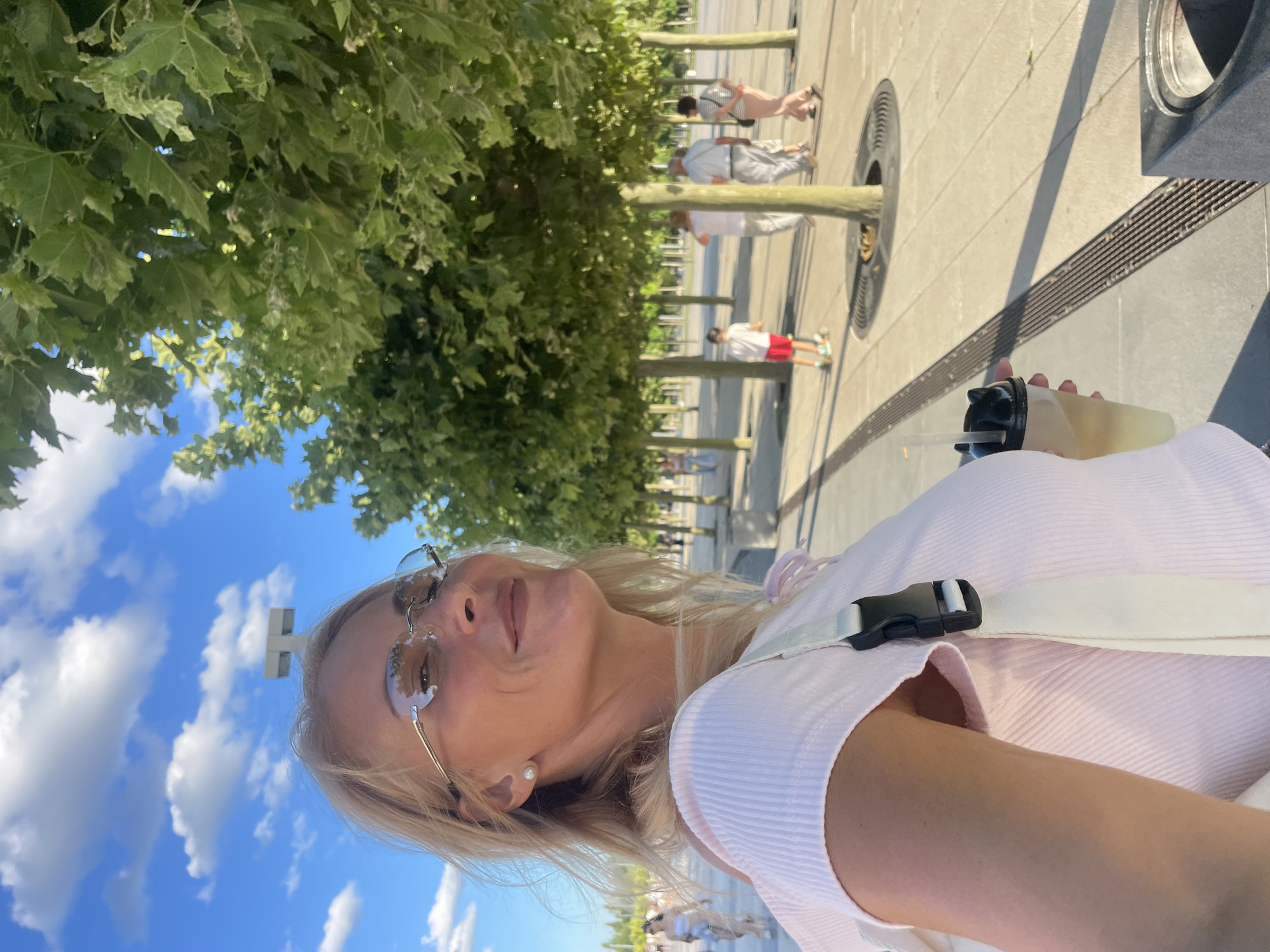 The image shows a blonde woman in her late 40s or early 50s, wearing a pink ribbed top and rimless glasses, taking a selfie outdoors. She is holding a drink with a cat-shaped lid and a straw. In the background, a paved walkway lined with trees can be seen. Several people are walking in the distance, including a child on a scooter. The sky is blue with scattered clouds. The location is likely the Krasnodar City Park in Krasnodar, Russia, given the presence of organized green spaces with paved walkways and trees, and the general urban setting. The time of day appears to be late afternoon or early evening due to the lighting and long shadows.