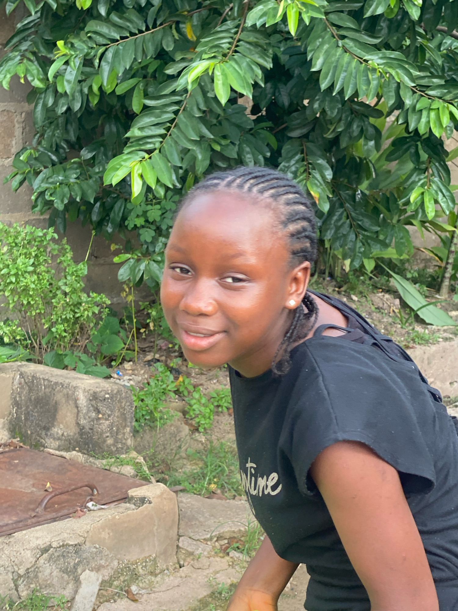 A medium shot captures a young Black female with braided hair and a black t-shirt from a slightly high angle, positioned in an outdoor, natural environment. She is looking toward the right of the frame with a slight smile. Her t-shirt has white writing on it. The background consists of green foliage and a brick wall. The foreground shows concrete structures and some greenery. The setting appears to be residential, possibly in Sabon Tasha, Nigeria.