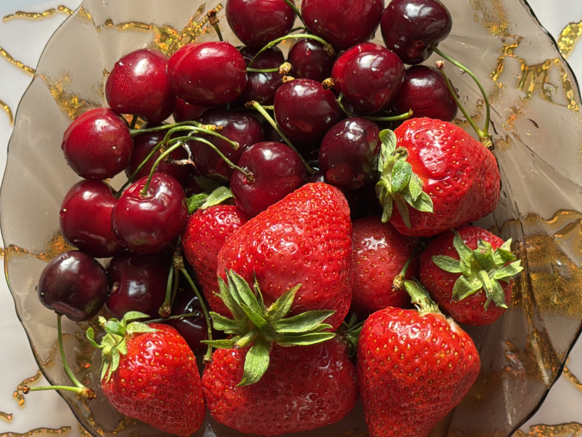A close-up, top-down shot shows a decorative glass bowl filled with fresh strawberries and dark red cherries. The bowl has a wavy rim and a textured, reflective gold pattern on its surface, set against a white, textured background. The fruits are densely packed, with some cherries obscuring strawberries and vice versa. The strawberries, predominantly in the foreground and lower portion of the image, are bright red with visible seeds and green leafy tops. The cherries are deep red to almost black, some appearing to have water droplets on their skins. The lighting casts subtle shadows and highlights, emphasizing the glossy texture of the fruits and the metallic sheen of the bowl. No people or actions are depicted; it is a still life composition of fruit in a bowl.