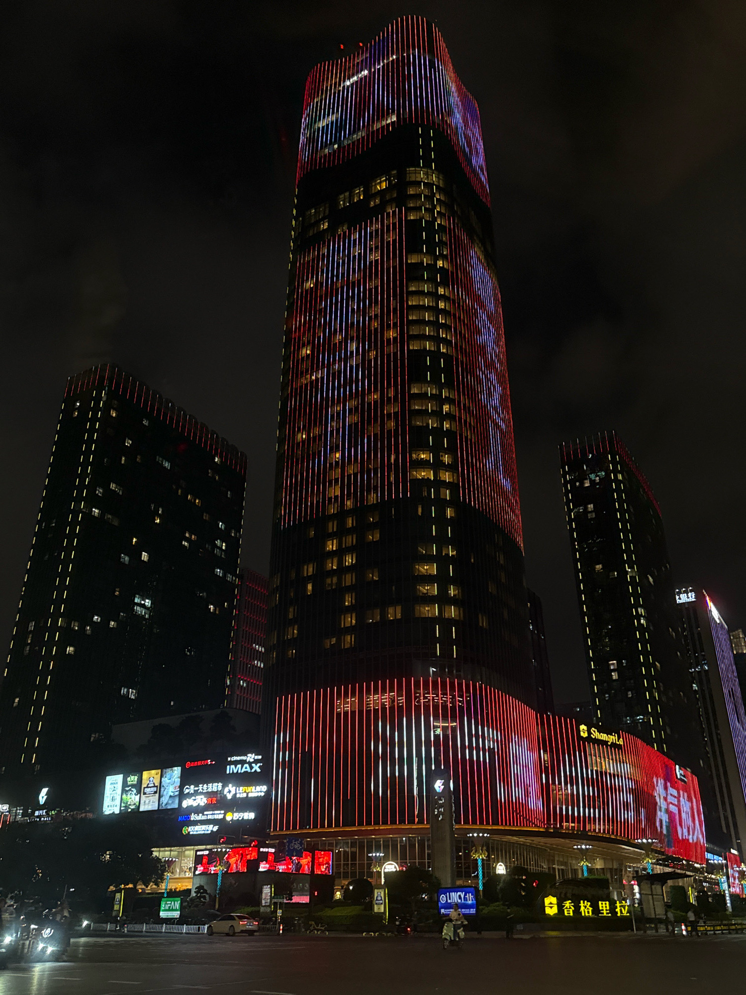 The image captures a nighttime view of a modern city street scene in Yiwu, China, featuring towering skyscrapers illuminated by vibrant LED lights. The central focus is a tall skyscraper adorned with vertical lines of red and white lights, creating a striking visual display against the dark sky. To its left and right are other buildings, also lit up, though with less elaborate patterns. The lower portion of the central skyscraper and adjacent buildings showcases a shopping complex or entertainment venue with large illuminated signs, including one that appears to say "Shangri-La" and another with Chinese characters and what seems to be an advertisement for a cinema with "IMAX" prominently displayed. A street with vehicles, including cars and a scooter, is visible in the foreground, suggesting an active urban environment. The overall impression is one of a bustling commercial district at night, characterized by impressive architecture and a lively atmosphere.