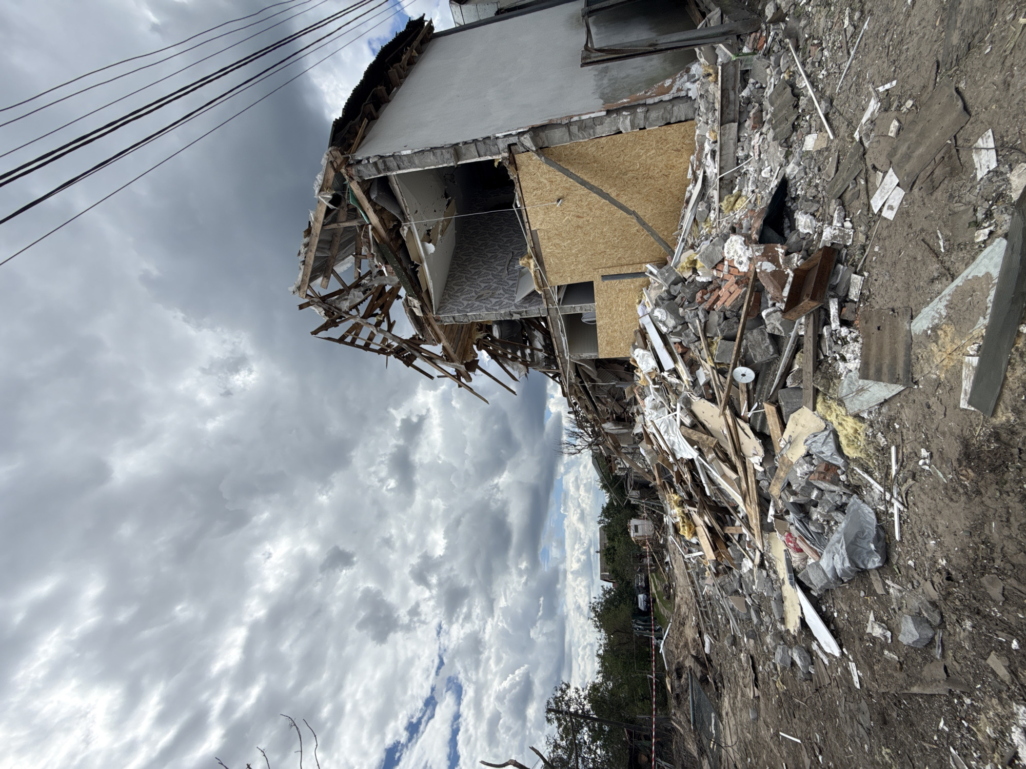 The image depicts the aftermath of destruction in Kyiv, Ukraine, with a heavily damaged multi-story building as the central subject. The building's facade is severely compromised, revealing exposed interior walls with patterned wallpaper and structural wooden beams. Debris, including concrete, wood, brick, and various construction materials, is scattered extensively around the base of the building and stretches into the foreground, forming a large pile. In the background, trees and other structures are visible, suggesting a residential or mixed-use area. Power lines stretch across the sky, which is filled with overcast, dark clouds. The scene conveys a sense of significant destruction and devastation. There are no people visible in the image.