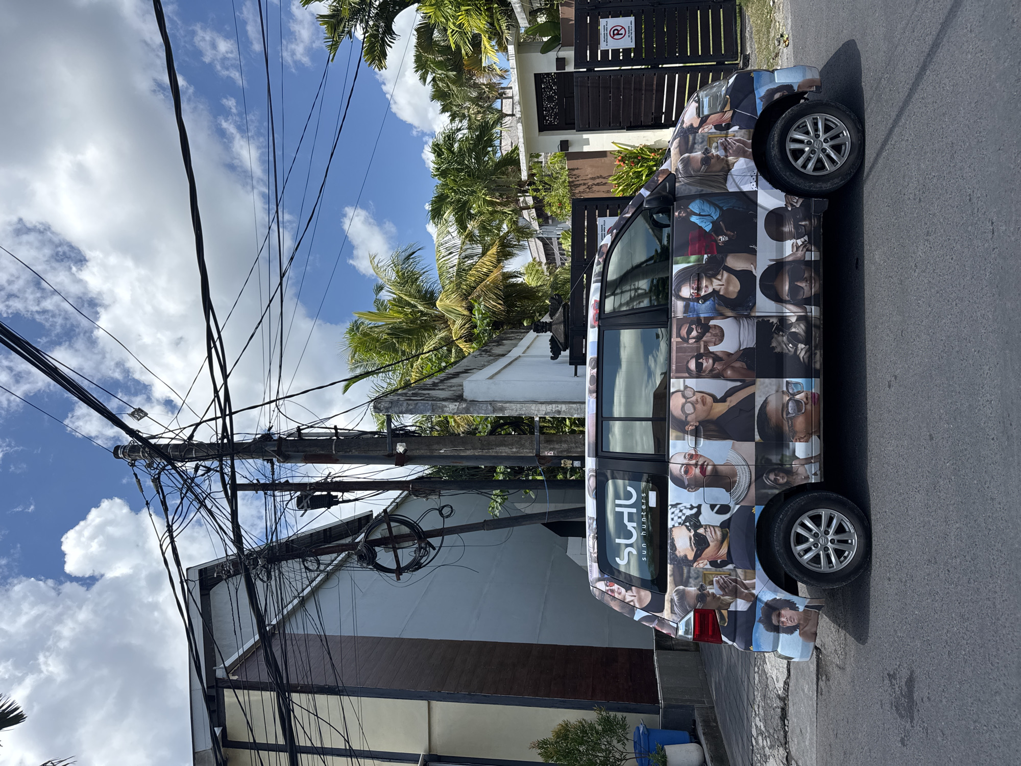 The image presents a street scene in North Kuta, Indonesia, featuring a silver minivan with a distinctive advertisement wrap displaying a grid of individual portraits. The minivan is parked on the side of a paved road, with the rear of the vehicle closest to the viewer.
Behind the minivan is a low-rise building, primarily white with a horizontal brown accent band. Adjacent to the building is a utility pole densely laden with electrical wires. The sky above is partly cloudy, with blue patches visible between the clouds.
In the background, there's a glimpse of tropical vegetation, including palm trees. A gate and partial view of another building can also be seen beyond the trees. The overall impression is that of a residential or mixed-use neighborhood street scene on a sunny day in a tropical location.