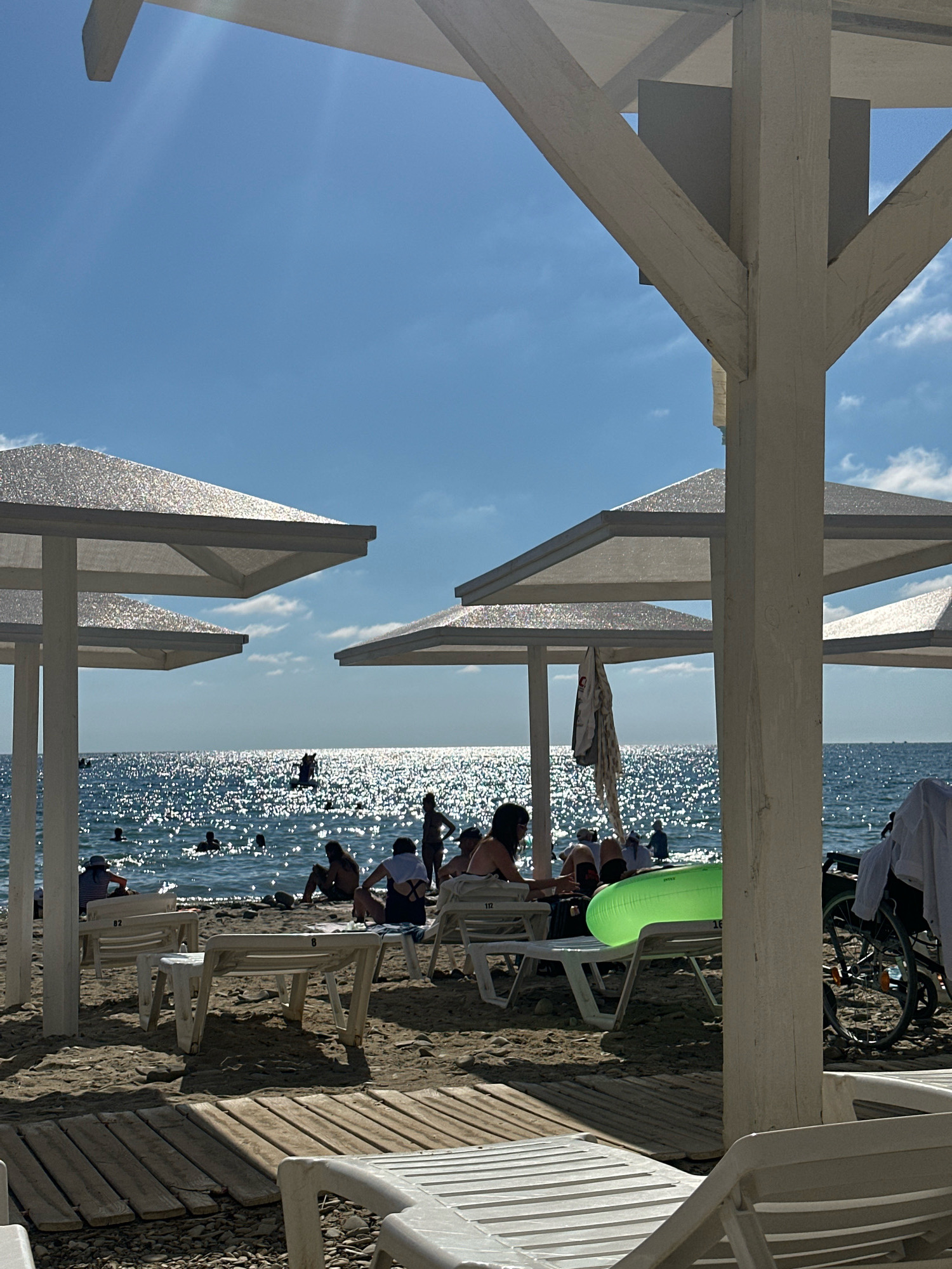 The image depicts a sunny day at a beach, likely in Lermontovo, Russia, given the architectural style of the beach structures. In the foreground, several white plastic lounge chairs are arranged on a sandy and pebbly beach. Wooden planks are laid out as pathways. To the left and center, white canopies supported by white wooden posts offer shade, with similar structures visible further back.
The sea is visible in the background, sparkling under the bright sun with numerous small waves. Several people are seen in the water, some swimming, others standing. Silhouettes of beachgoers are also visible along the shoreline and seated on lounge chairs under the canopies. One person is on a floating device in the water.
A green inflatable ring rests on a lounge chair. To the far right, a wheelchair is parked next to a white wooden post. A piece of white clothing is draped over a wooden beam of the canopy structure. The sky is blue with scattered white clouds. The overall scene conveys a relaxed atmosphere of a summer day at the seaside.