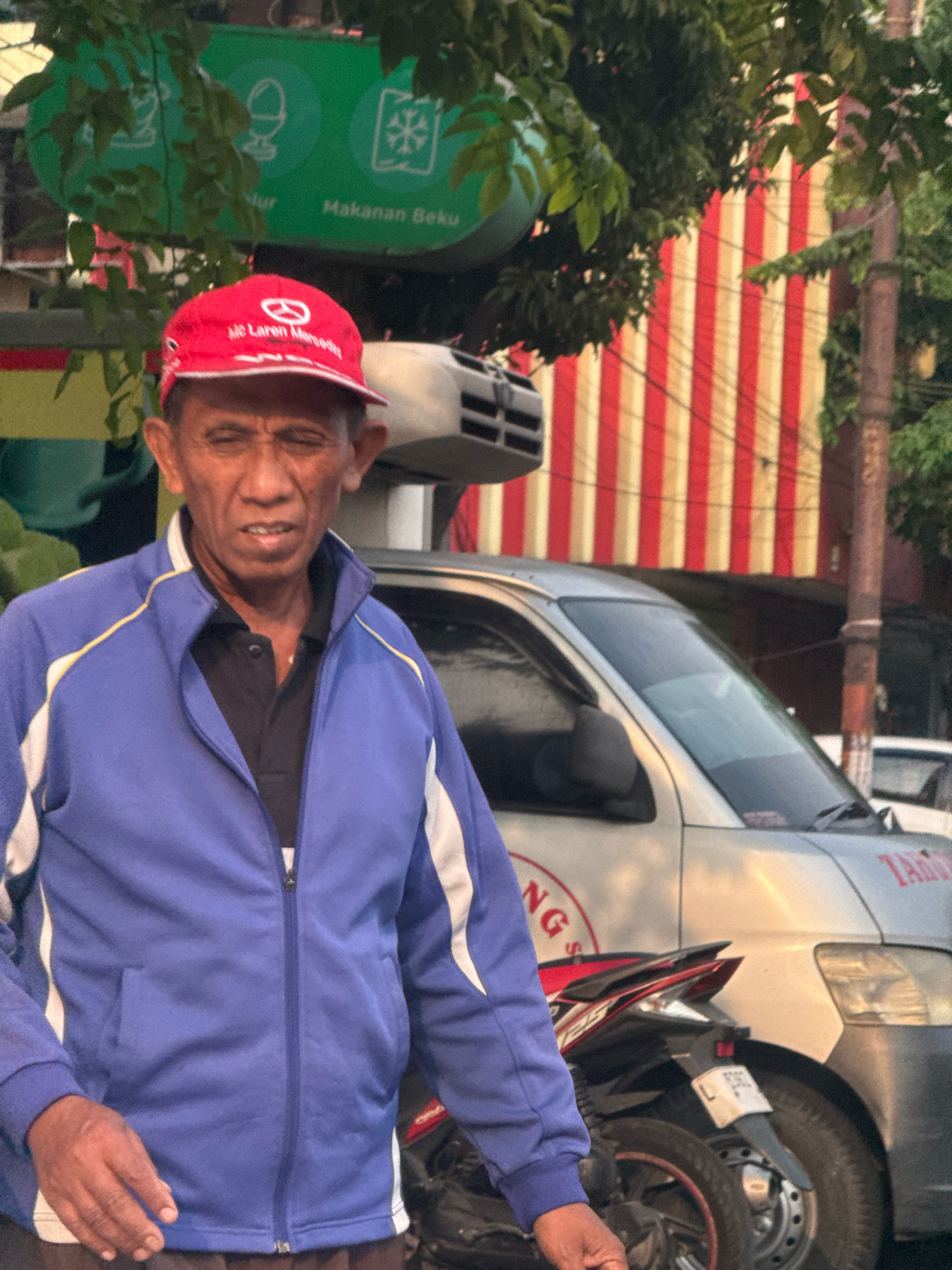 An older man with dark skin and dark hair, wearing a red baseball cap and a blue zippered jacket over a black shirt, stands in front of a silver van and a motorcycle. The man's mouth is slightly open, and his eyes are squinted. The van has red and white striped banners behind it, and what appears to be a logo on its side. A motorcycle is parked to the right of the man. The scene appears to be in an urban environment, likely in Surabaya, Indonesia, given the presence of the man and the style of the van. Green foliage is visible at the top of the frame.
