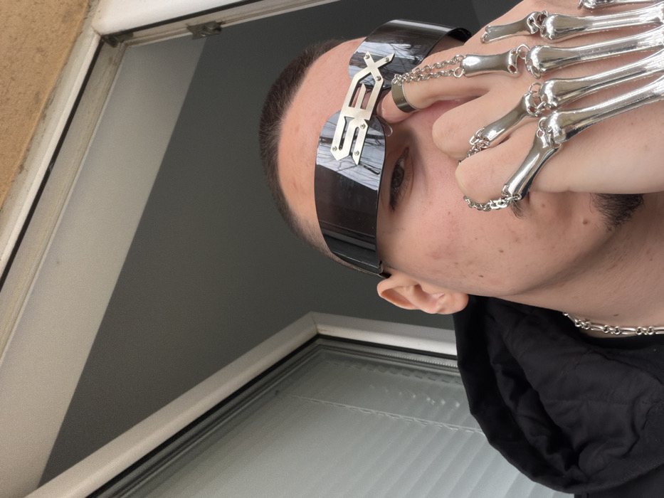 A male individual is depicted in a low-angle, head-and-shoulders shot within the city of Varna, Bulgaria. The subject wears dark, reflective, wraparound visor-style sunglasses prominently featuring the word "SEX" in metallic silver letters across the bridge. His hair is closely cropped, and a goatee is visible. His left hand is raised, positioned near his lower face and nose, adorned with five articulated, silver-colored skeletal finger extensions connected by thin chains to a plain silver band ring on his left index finger. He is wearing a dark-colored hooded garment and a silver-colored chain necklace. The background shows a white-framed window with visible internal blinds on the left, and a light-colored, textured building surface or ceiling on the upper right. The subject is looking directly at the camera.