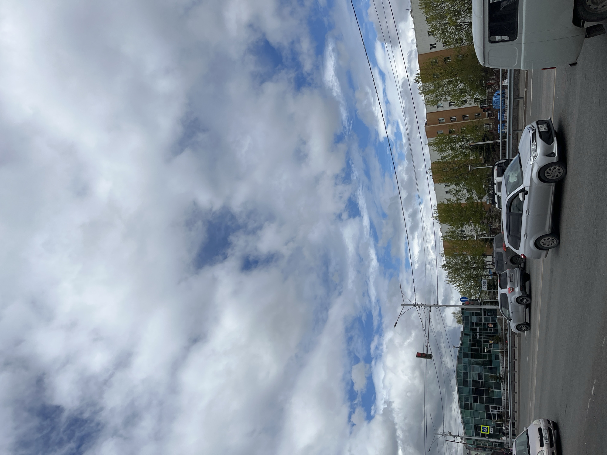 An eye-level, medium-shot, vertical photograph captures a street scene in Petropavlovsk-Kamchatsky, Russia, under a dramatic sky filled with billowing clouds. In the foreground, asphalt pavement dominates, with several vehicles in motion. A silver sedan is on the left, while a silver SUV navigates the intersection in the center. To the right, the rear of a white van is visible.
The midground features a modern, glass-fronted building on the left, reflecting the sky and surrounding environment. Traffic lights and street lamps are visible, indicating an urban intersection. Across the street, a long, multi-story building with a patterned facade is lined with trees. The street is bordered by a metal railing.
The background is dominated by the expansive sky, characterized by a mix of dark and light clouds, with patches of blue sky peeking through. The overall impression is of a cloudy day in a Russian city.