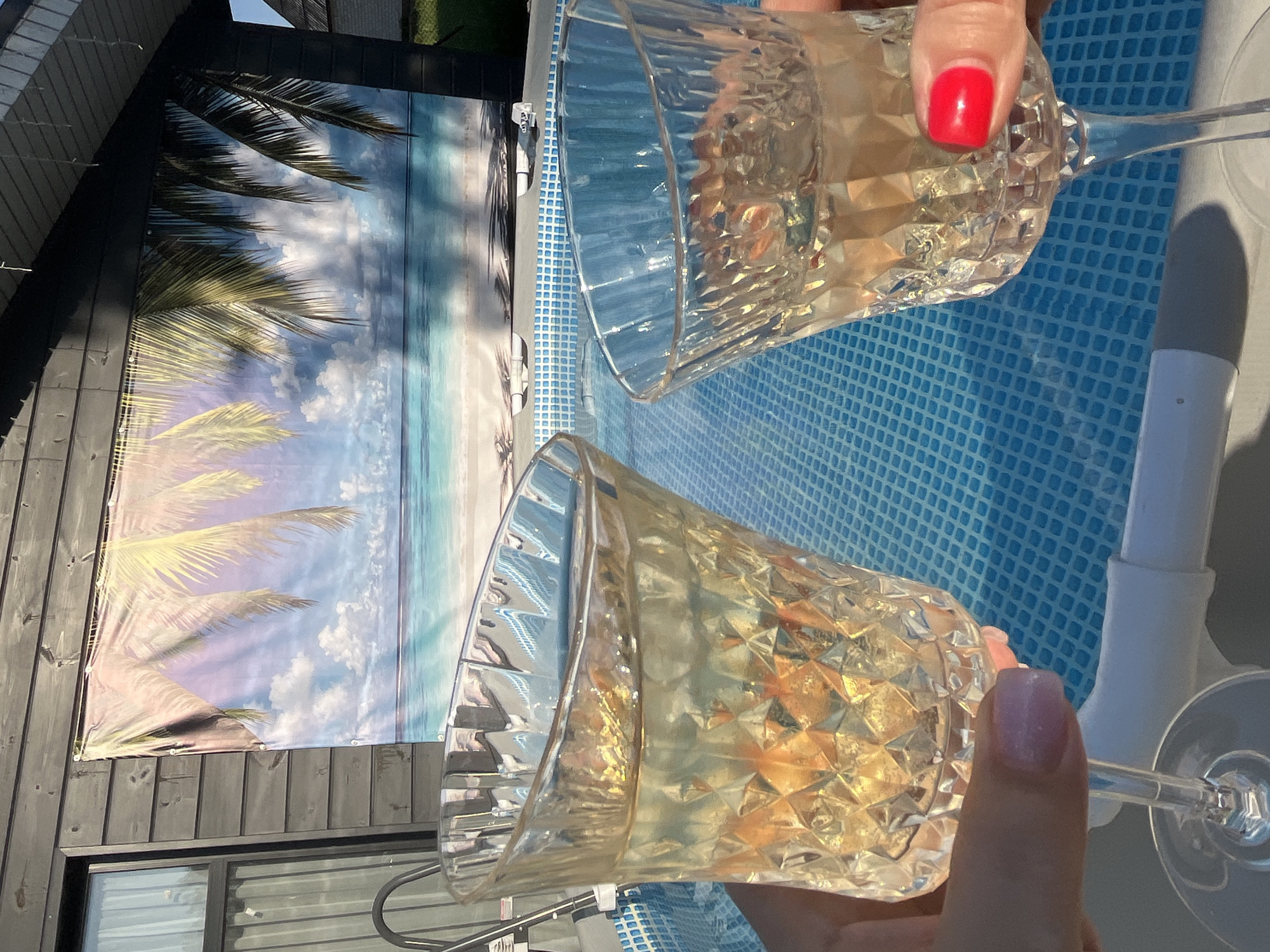 The image captures a celebration near a pool, possibly in Новая Чернушка, Russia. Two crystal glasses filled with a light-colored beverage are being clinked together in a toast. One hand has a red fingernail polish, while the other has a light pink polish. Behind the glasses, the pool's blue checkered pattern is visible. A backdrop depicting a beach scene with palm trees and an ocean hangs on a dark-colored wall, providing a tropical ambiance to the setting. A window is partially visible below the backdrop.