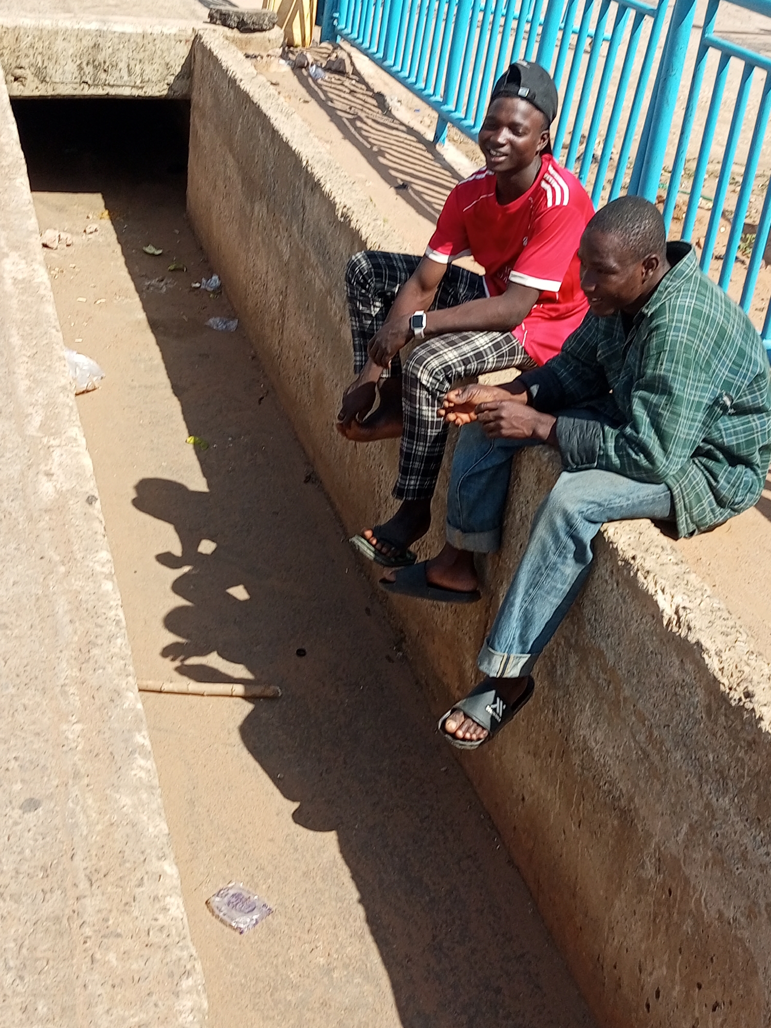 Two men are sitting on a concrete structure, likely a bridge or an overpass, in Garko, Nigeria. The man on the left is wearing a red t-shirt with white stripes on the sleeves, plaid pants, and a black baseball cap. He is smiling and looking to his left. The man on the right is wearing a green checkered shirt and blue jeans. He is looking forward. They appear to be engaged in conversation.
The surrounding environment is an open-air drainage or a dry water channel. The channel is made of concrete and is filled with sand and some debris. There are shadows cast on the ground, indicating a sunny day. In the background, there is a bright blue metal railing. The overall impression is one of a casual, everyday scene in an urban or semi-urban setting in Garko.