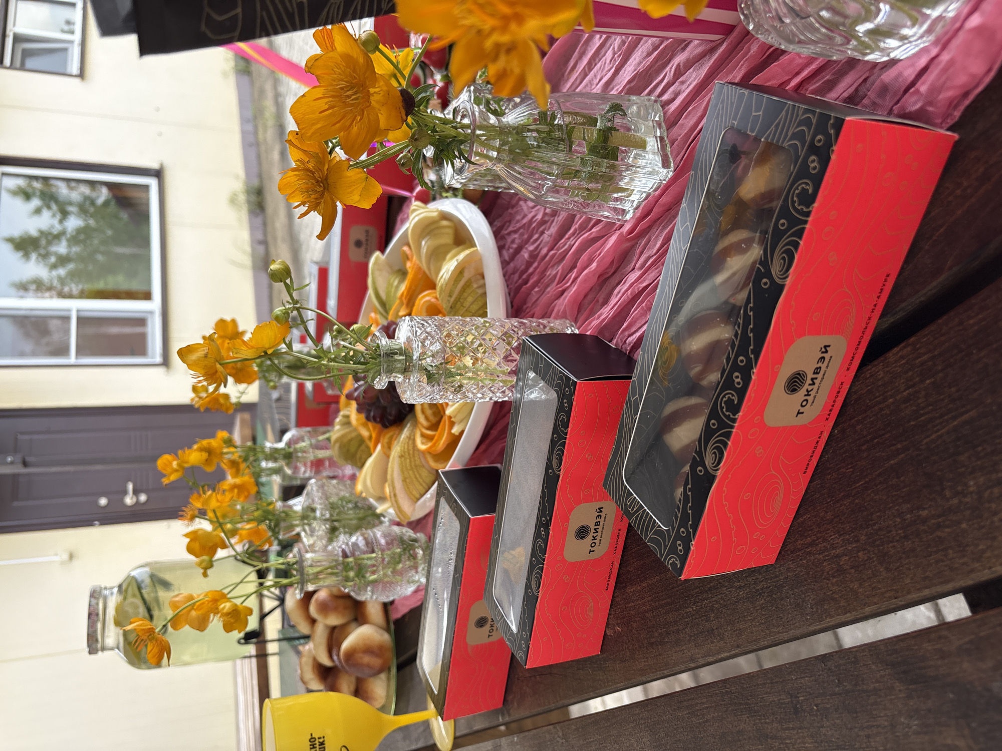 A wooden table is set with a spread of food and drinks, including pastries, fruit slices, and several boxes of food. There are also three glass vases holding small orange flowers. In the background, a building with windows and a door is visible. The scene appears to be outdoors, possibly in a backyard or patio area. The text on the boxes suggests the location is Birobidzhan, Russia.